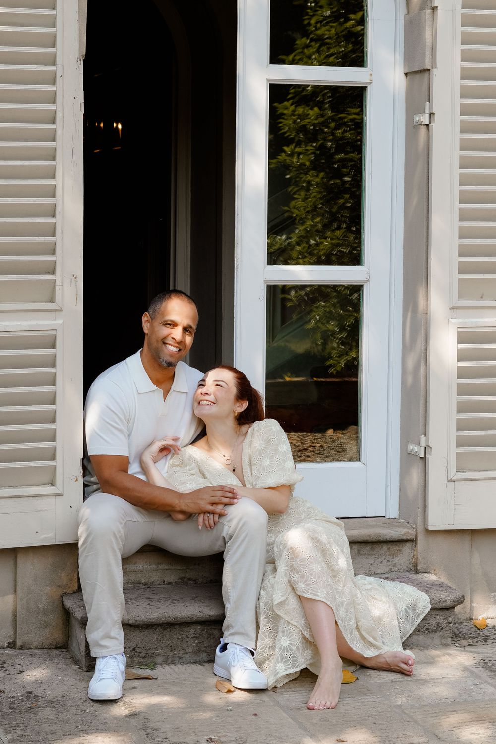 A happy interracial couple sits together on home steps, woman in floral dress leaning on man in white polo shirt.