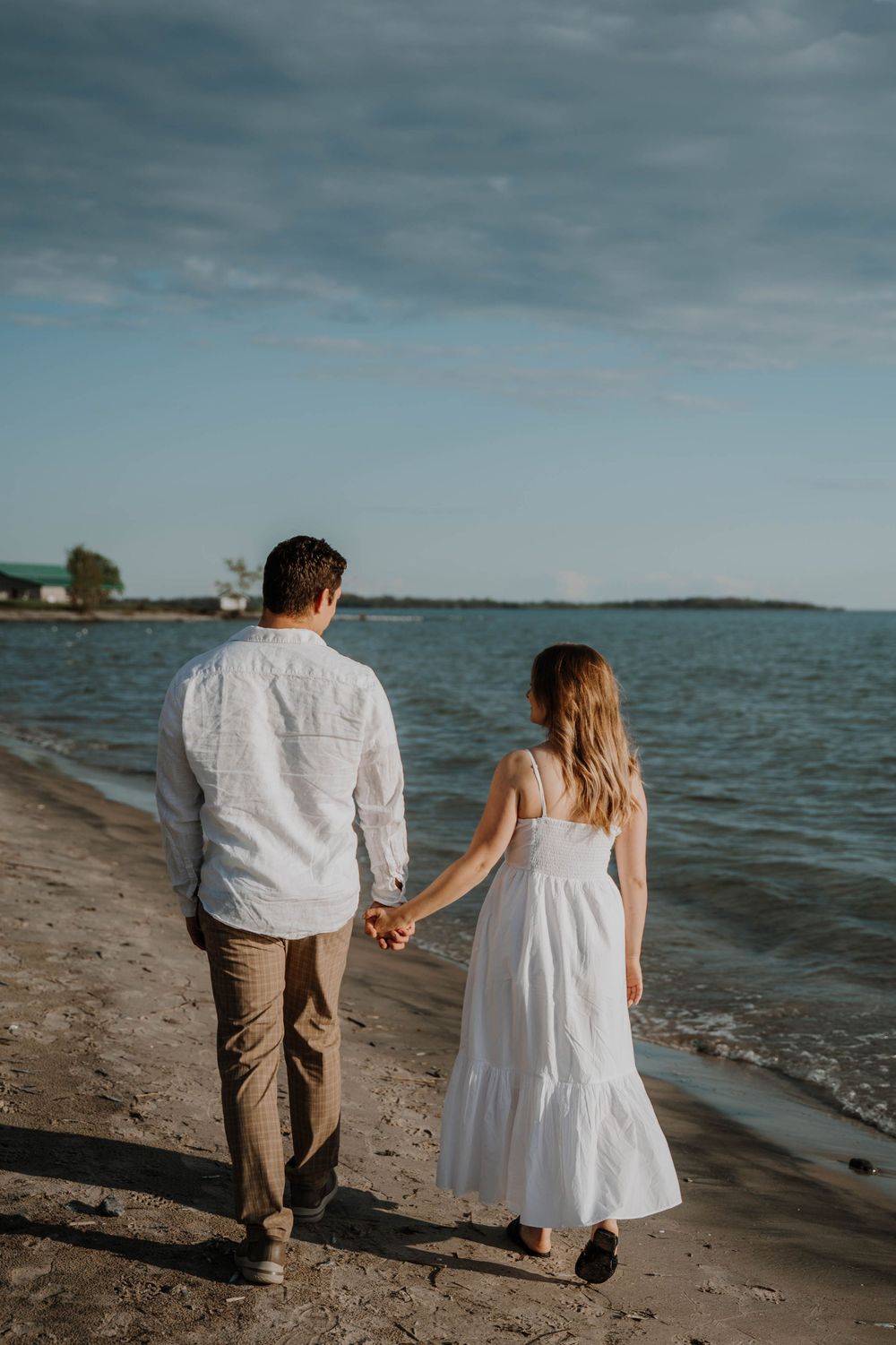A couple walks hand in hand along a serene beach shoreline at sunset, with the woman in white dress and man in casual attire.