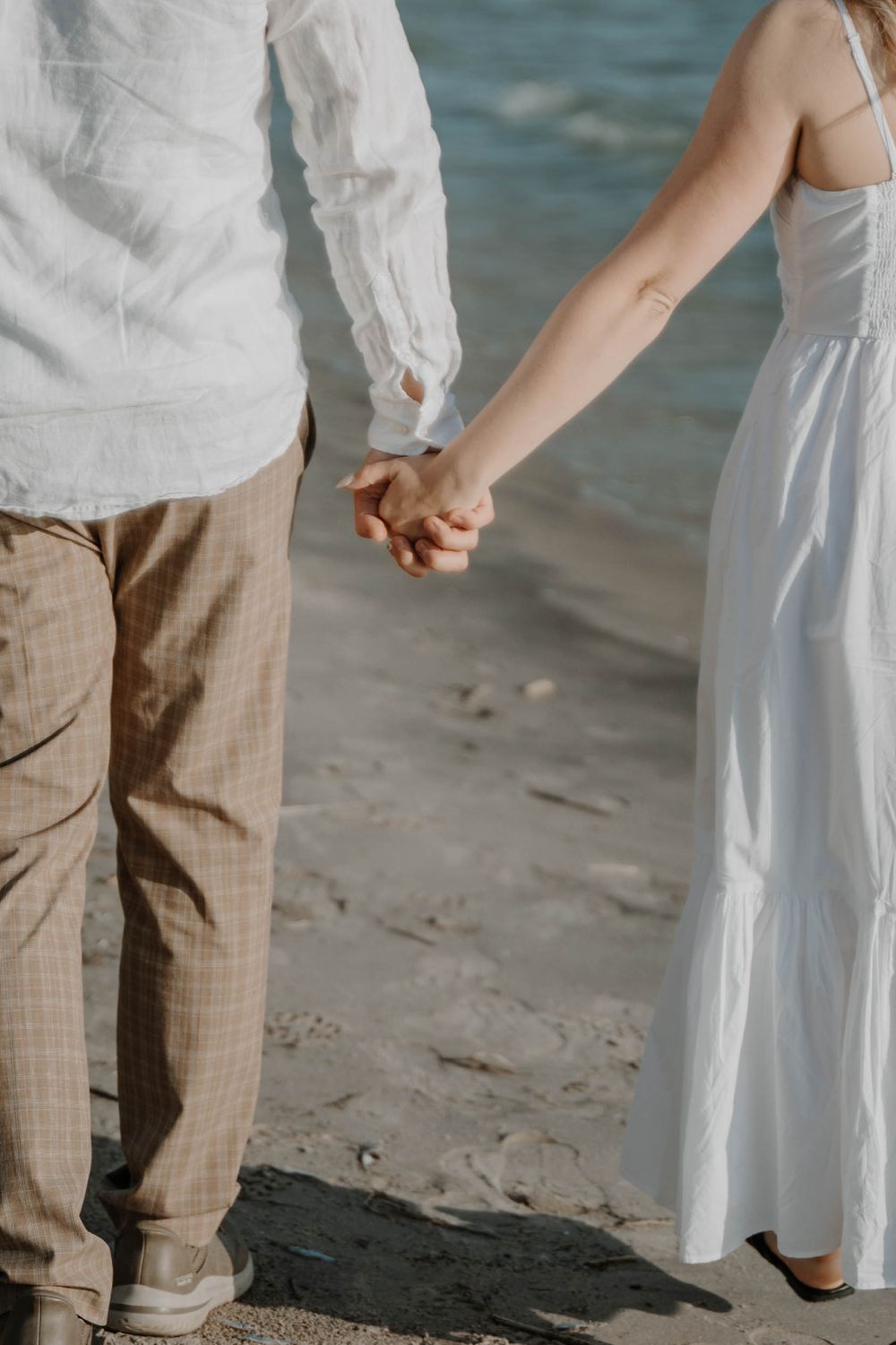 Close-up view of a couple holding hands while walking on a sandy beach with ocean waves visible in the background.