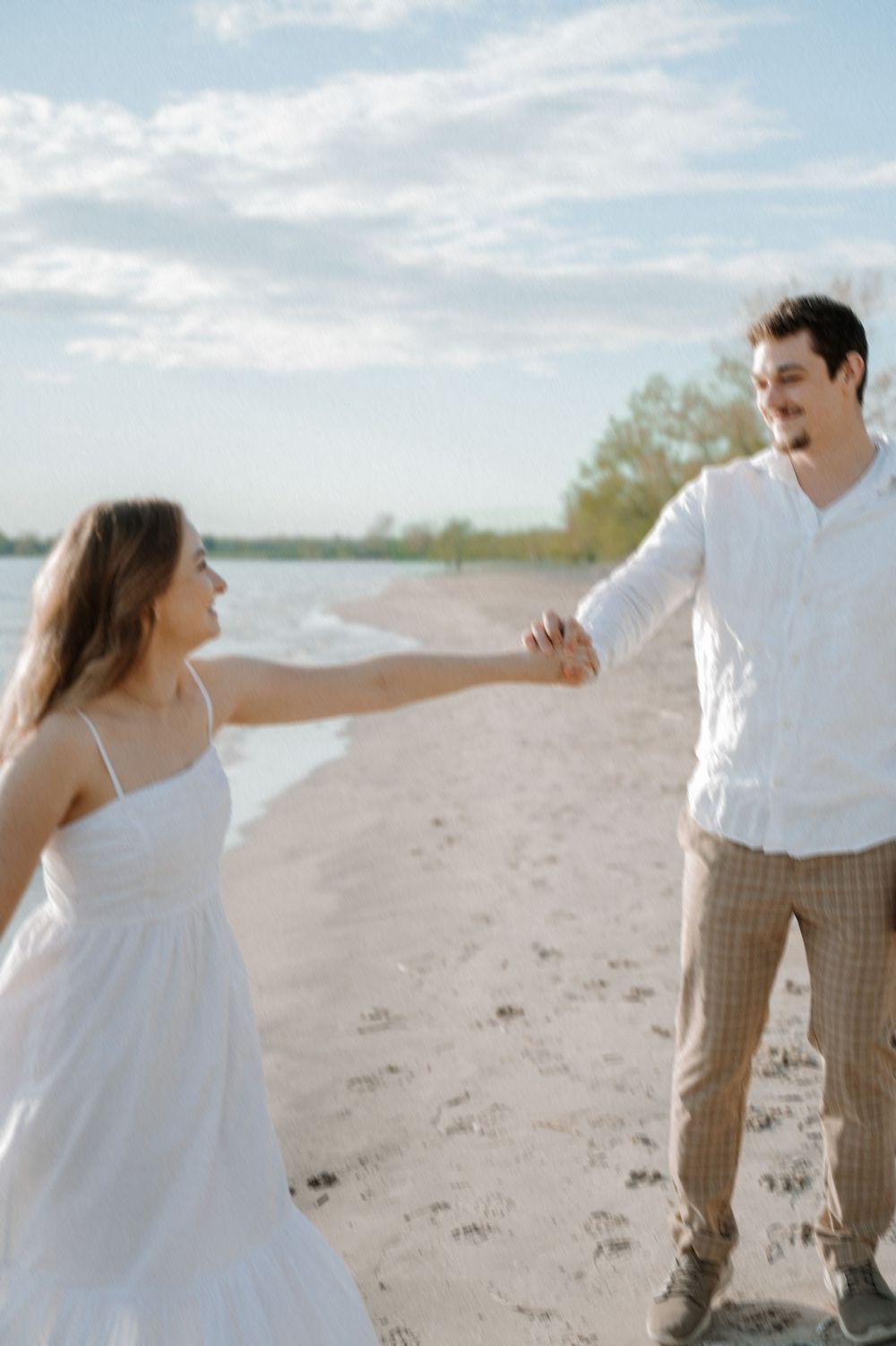A romantic moment captured on a sunny beach as a couple in white holds hands and smiles at each other.