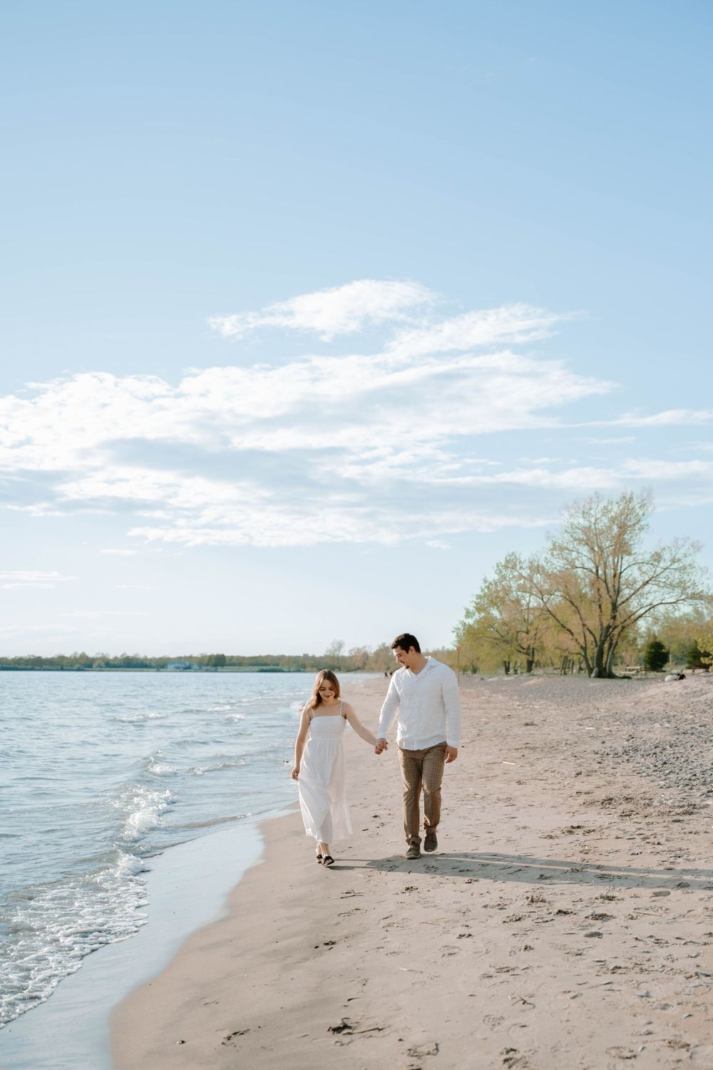 A couple strolls along a tranquil beach shoreline with water and clouds in the background on a sunny day.