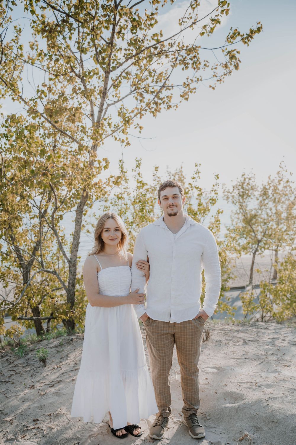 A casual engagement photo of a couple standing together outdoors with autumn foliage in the background.