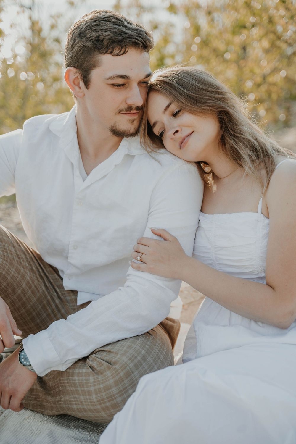 A romantic outdoor portrait of a couple in white clothing resting against each other with autumn colors in the background.