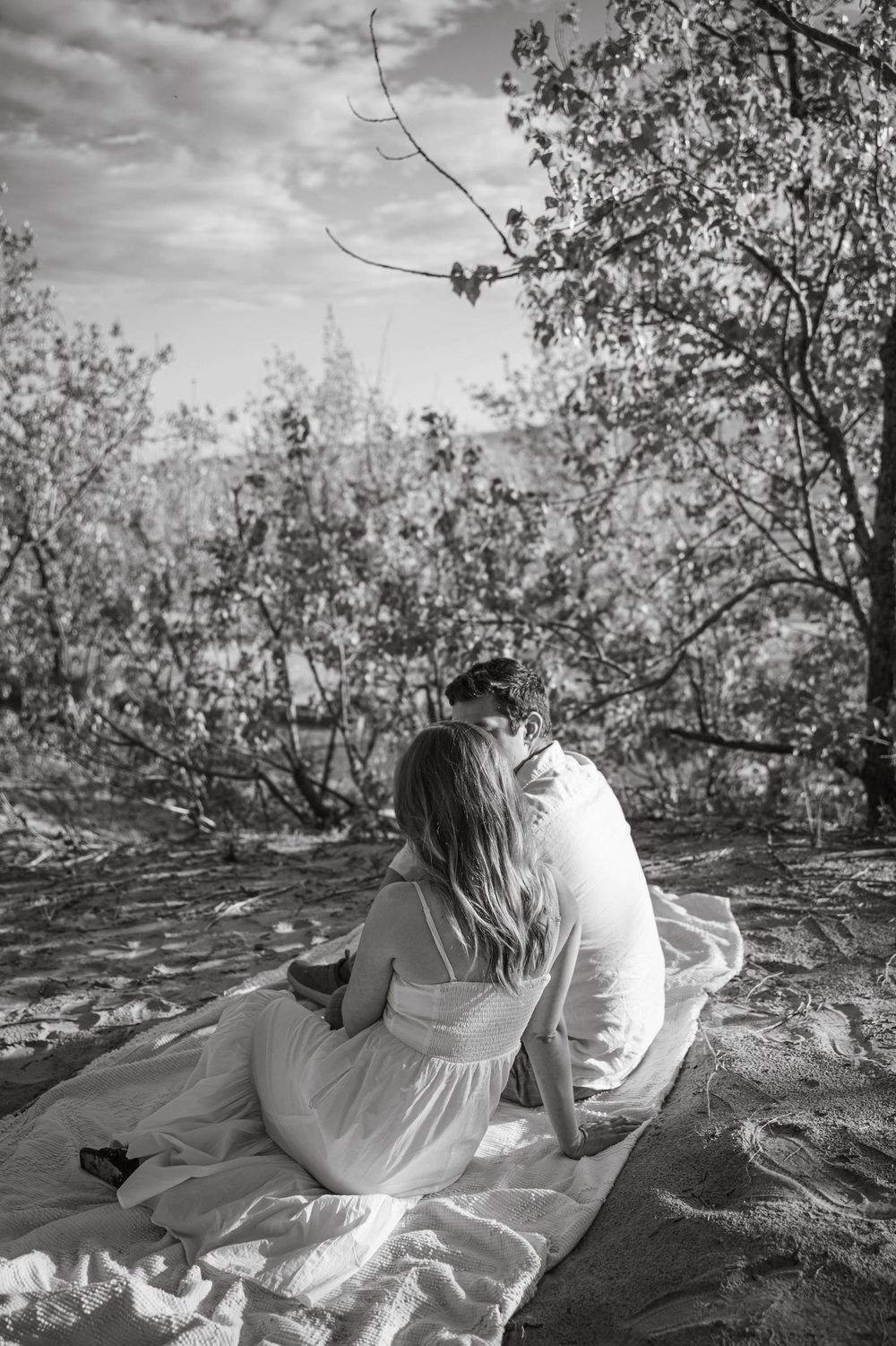 Romantic couple sitting on blanket at sunset surrounded by trees in black and white photography.