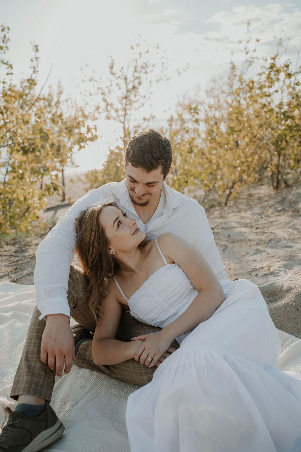 A romantic couple in wedding attire reclines in a sunlit natural setting with autumn foliage in the background.