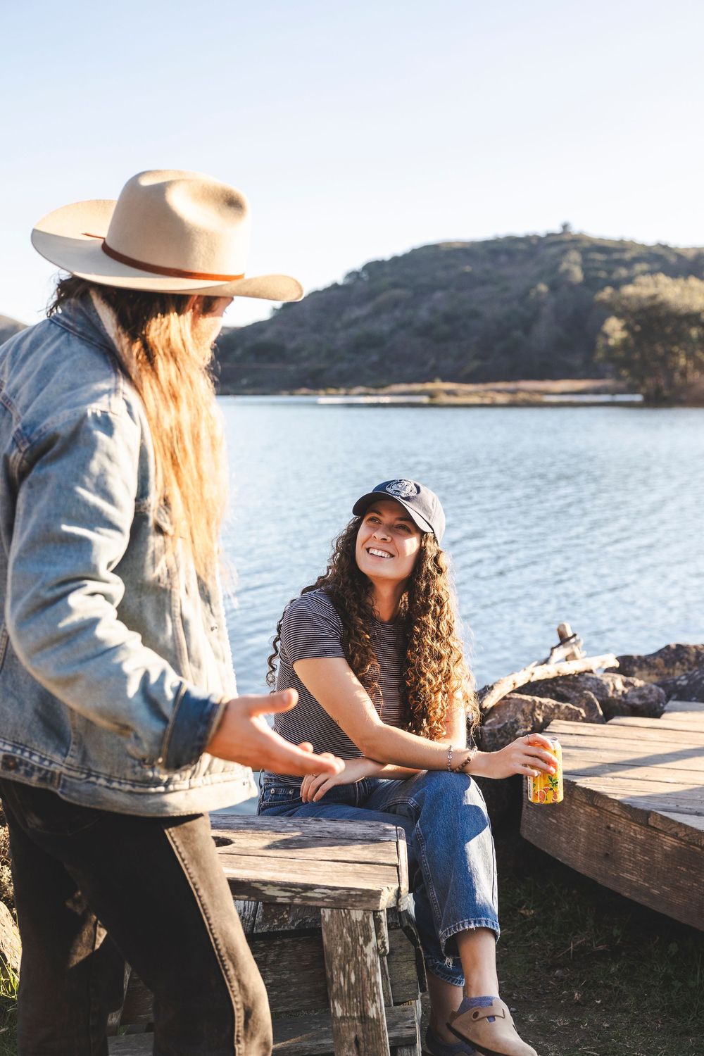 Friends enjoying drinks and conversation on a wooden dock by a scenic lake at sunset.