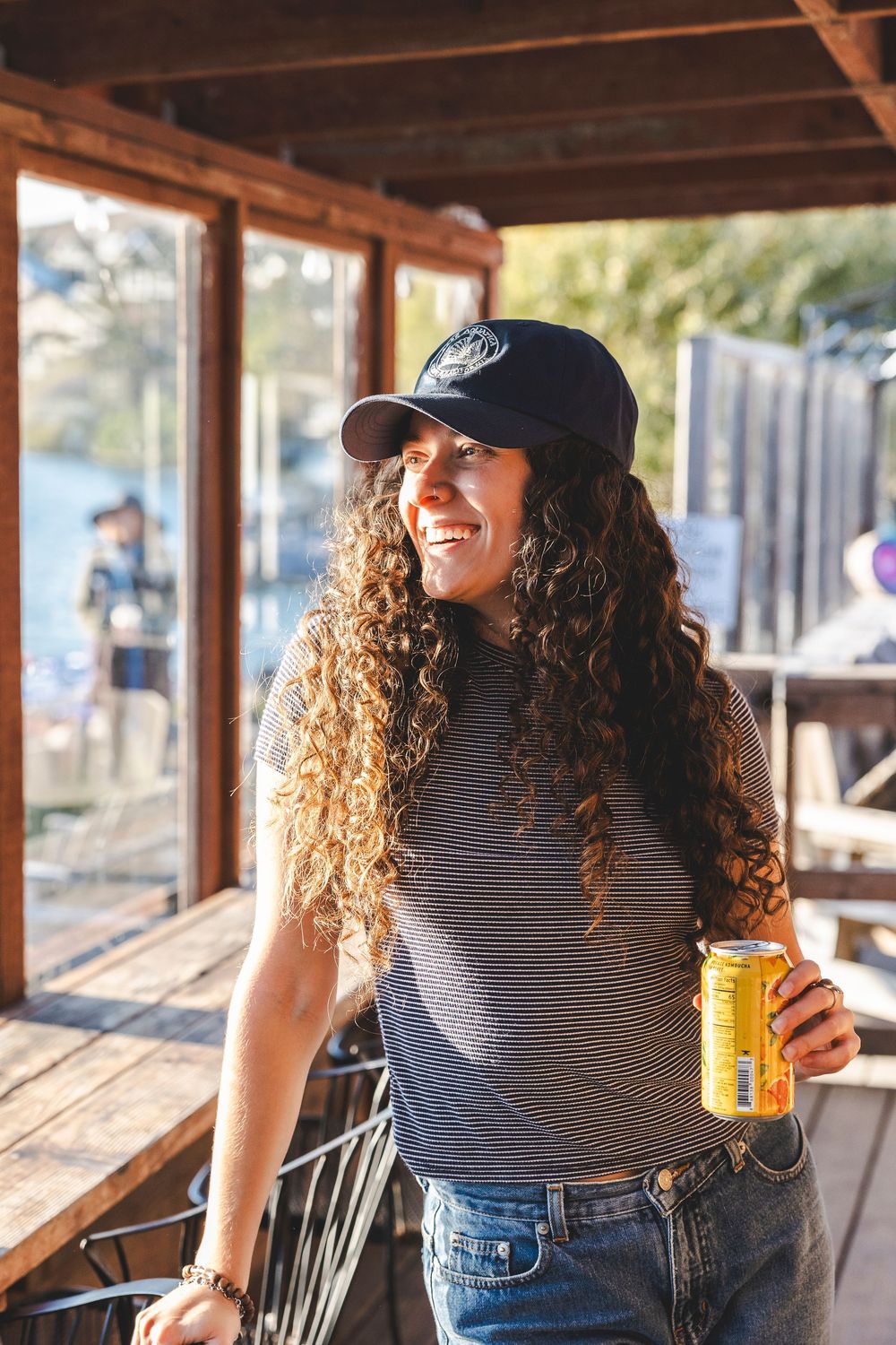 Person with curly hair wearing a striped shirt and baseball cap enjoys a drink on a wooden deck.