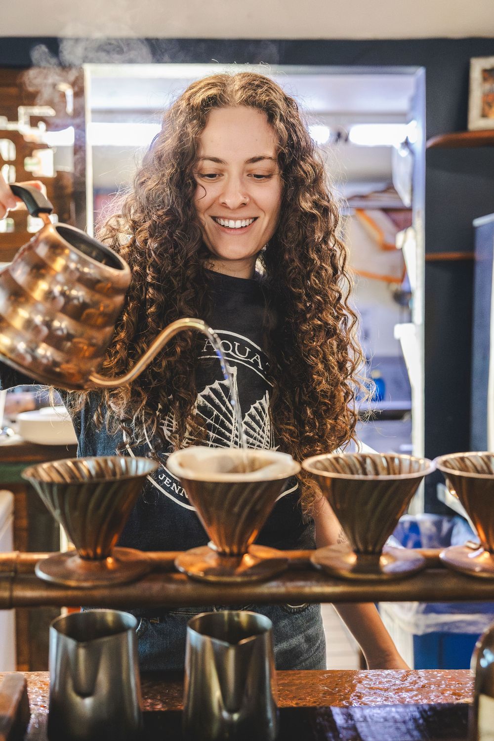 Barista with curly hair brewing coffee using multiple pour-over drippers in a cafe setting.
