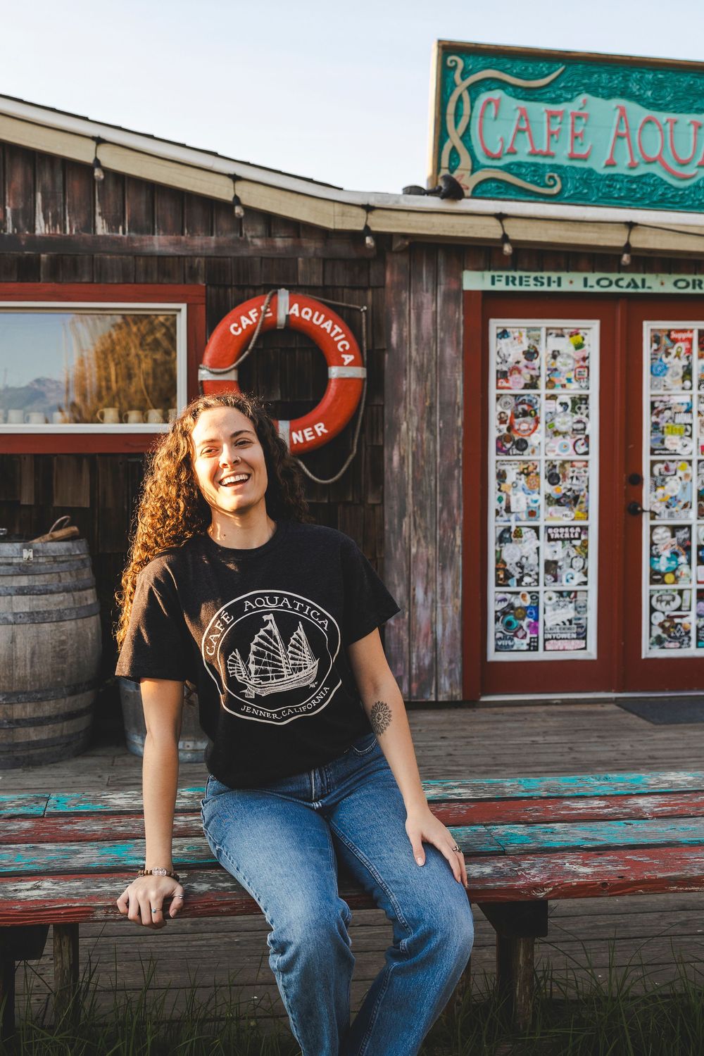 Person in black t-shirt and jeans sits casually on rustic wooden porch near vintage storefront.