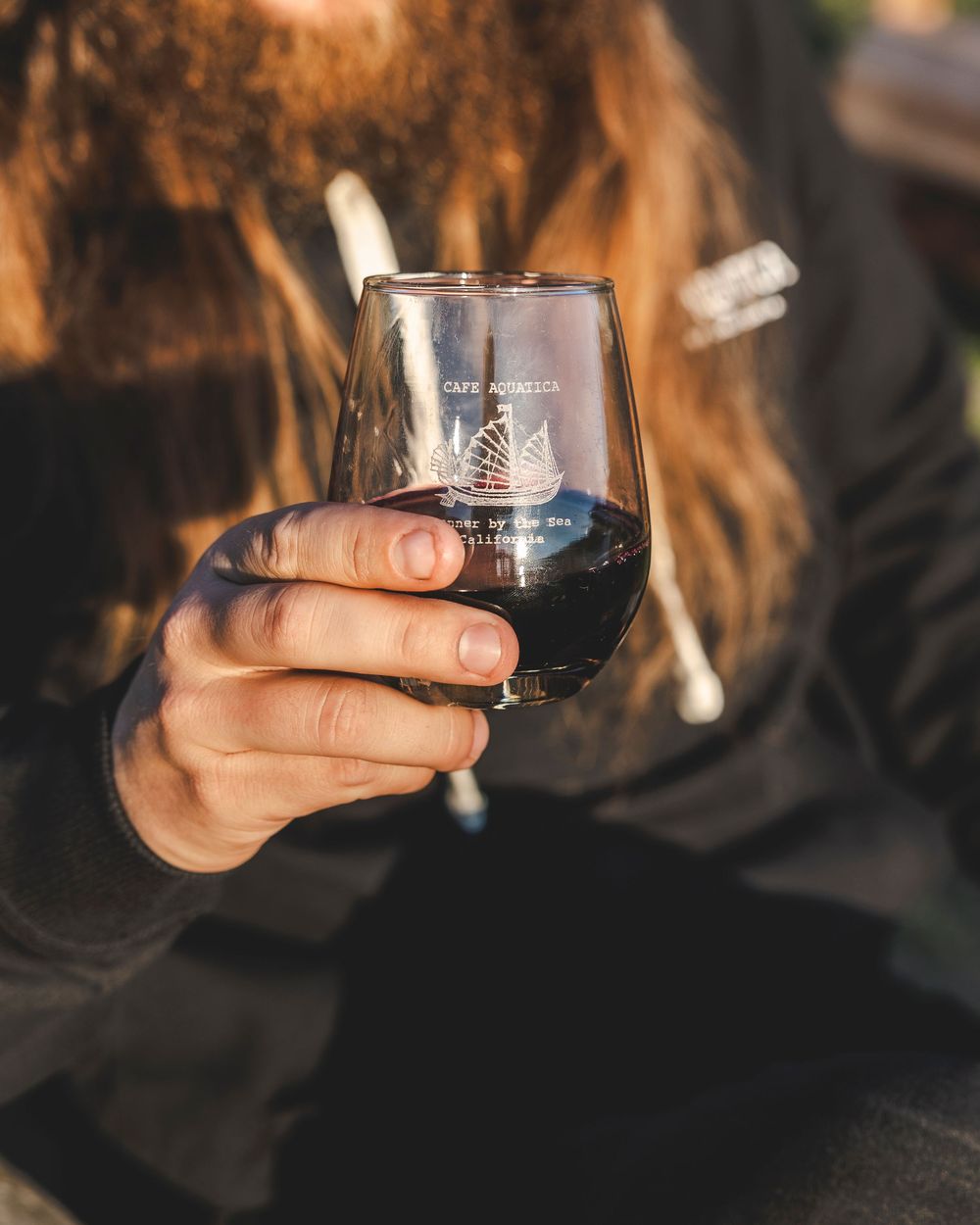 Hand holding a stemless wine glass filled with red wine against a warm sunset background.