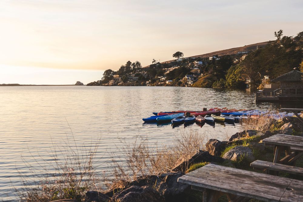 Row of colorful kayaks lined up along a serene lakeside shore at sunset with hillside homes in the distance.