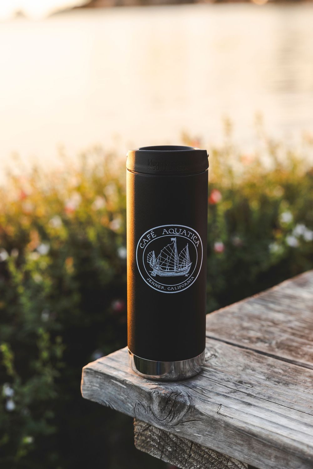 A black insulated drink holder with a boat logo sits on weathered wooden decking at sunset.