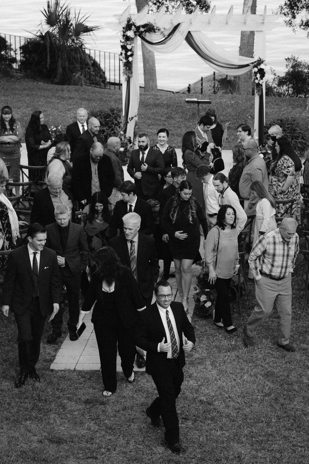 Black and white photo of wedding guests mingling outdoors near a floral arch after a ceremony.