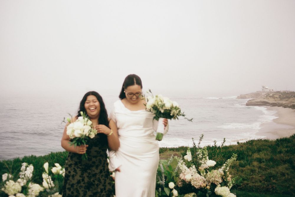 Bride in white gown holding bouquet with bridesmaid by coastal cliffside on foggy day.