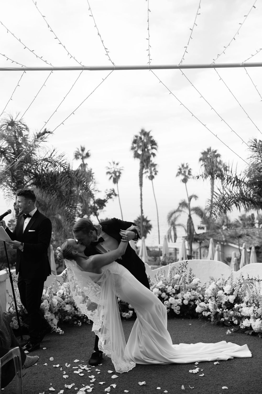 Bride and groom share romantic dip kiss during first dance at outdoor wedding reception with palm trees and string lights.