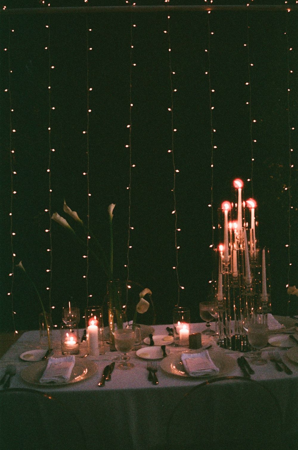 Romantic candlelit dinner table set for two with fairy lights backdrop and elegant candelabra centerpiece.