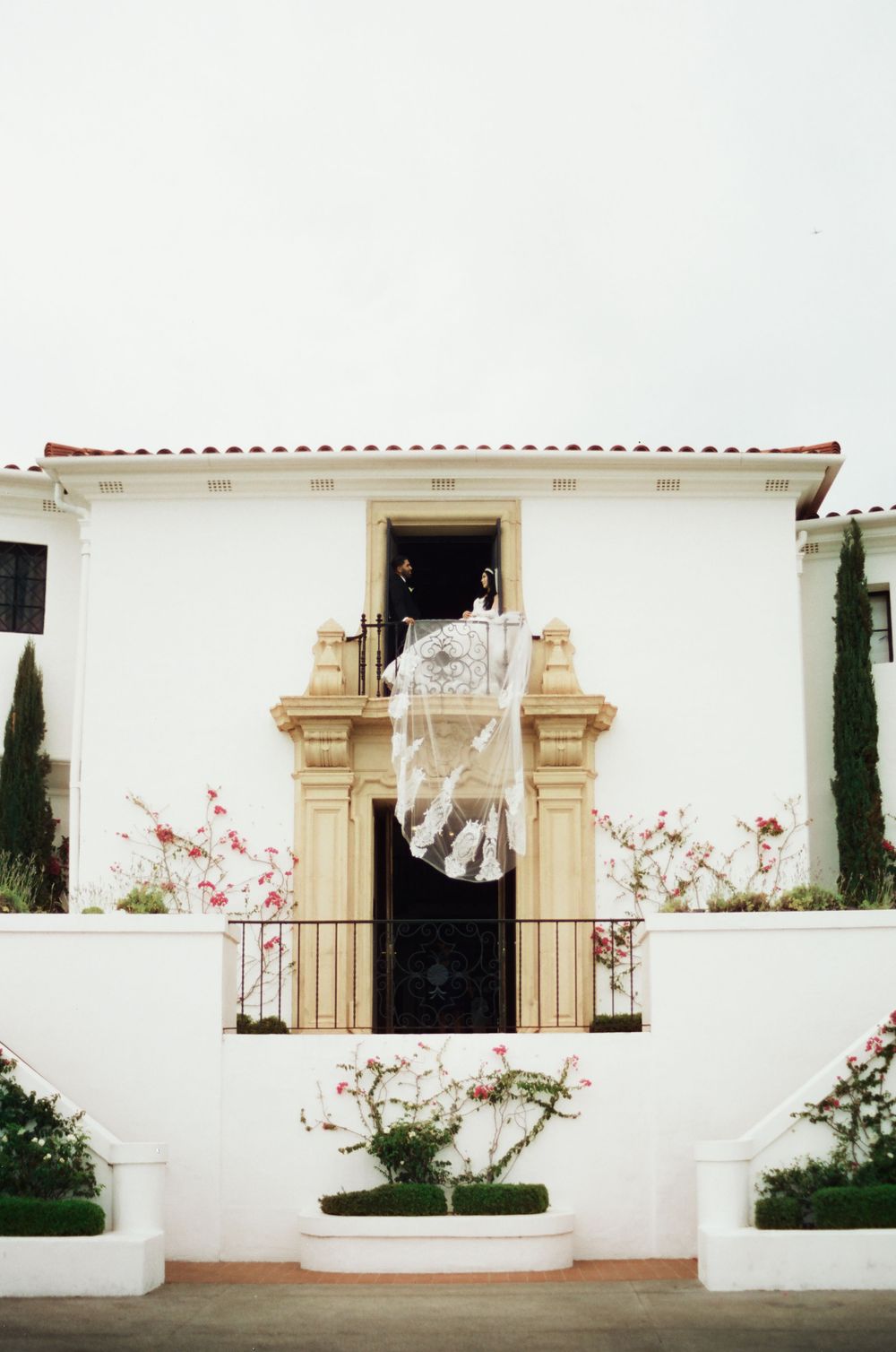 Bride displaying her lace wedding veil from an ornate balcony of a white Spanish-style wedding venue.