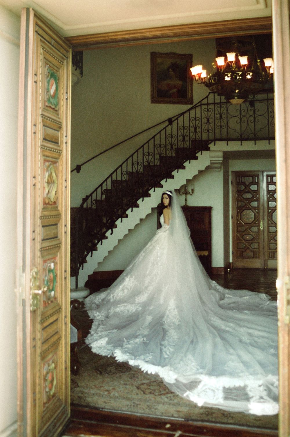 Bride in elegant white cathedral train gown poses in grand mansion foyer with ornate staircase and chandelier.