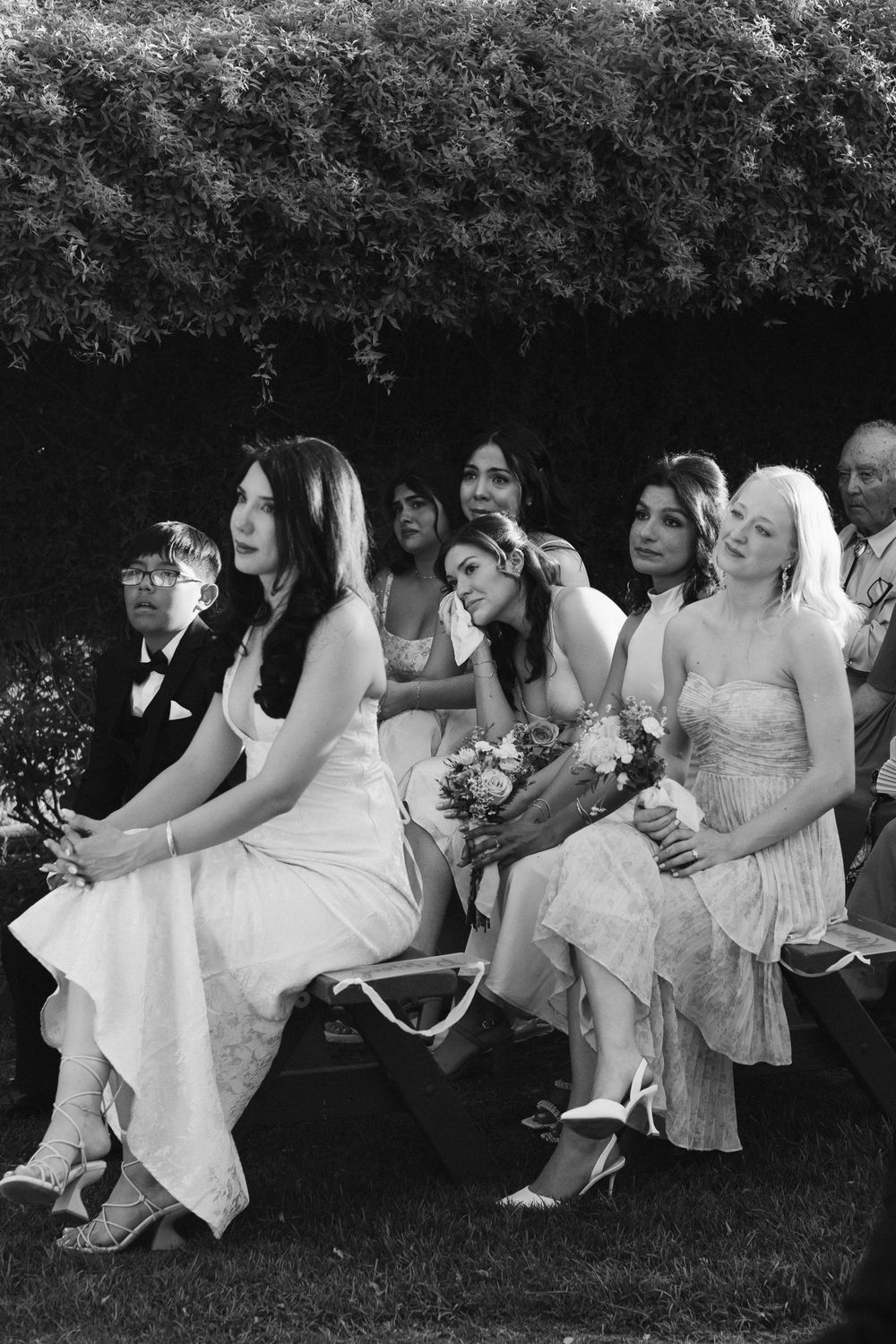 Black and white photo of bridesmaids seated outdoors at a wedding ceremony, some emotional, holding bouquets.