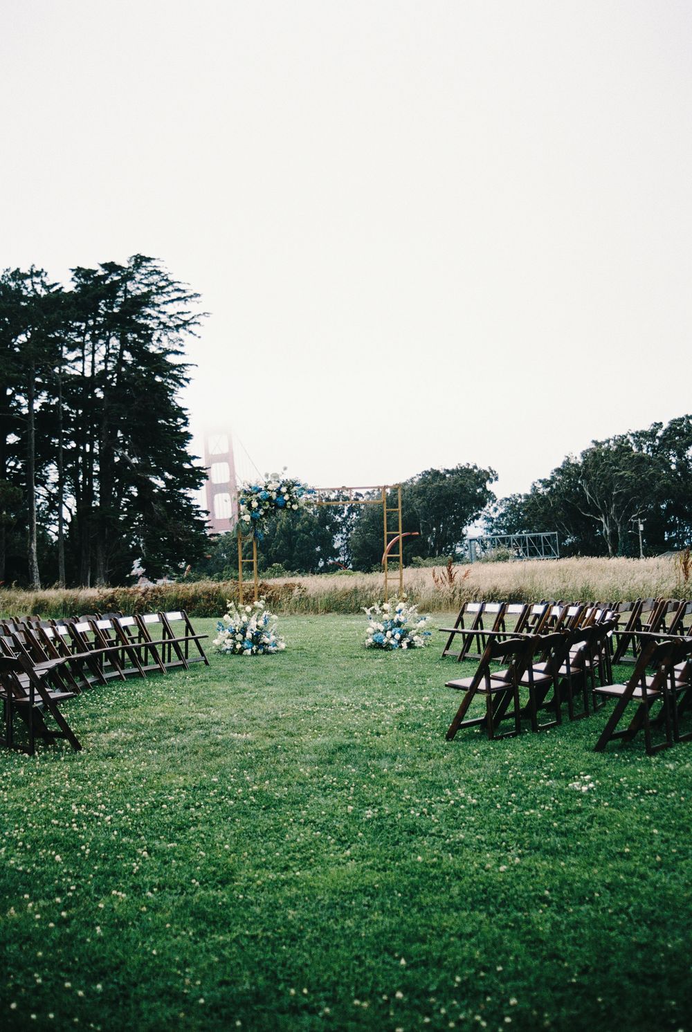 Outdoor wedding ceremony setup with wooden chairs on green grass, floral arrangements, and trees under an overcast sky.