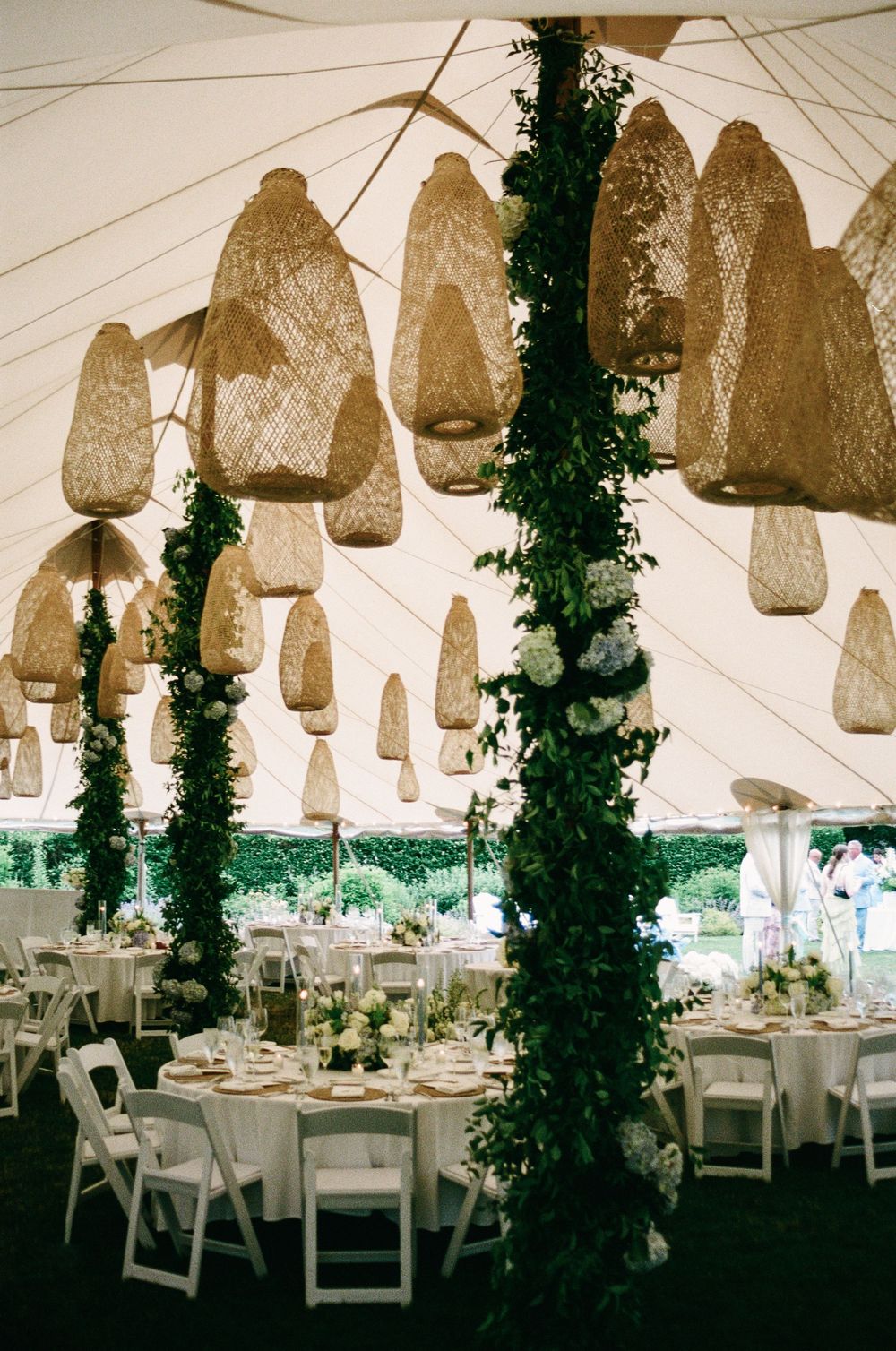 Elegant wedding tent reception with woven pendant lights, ivy-wrapped columns, white chairs, and floral centerpieces.