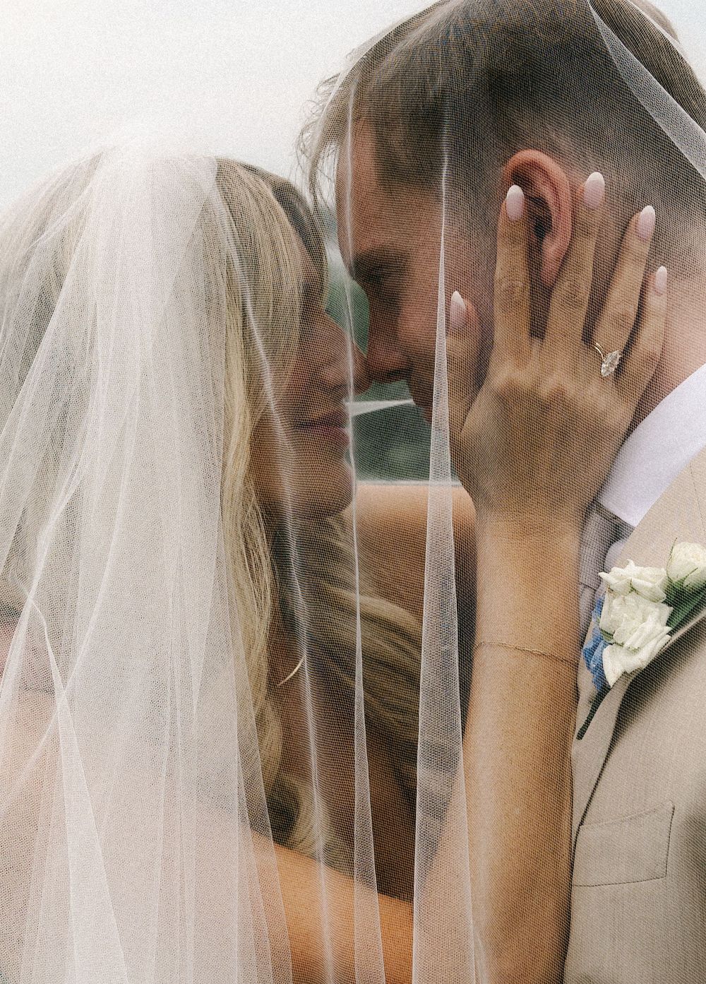 A bride and groom share an intimate moment under a flowing white veil, with the bride smiling and wearing a diamond ring.