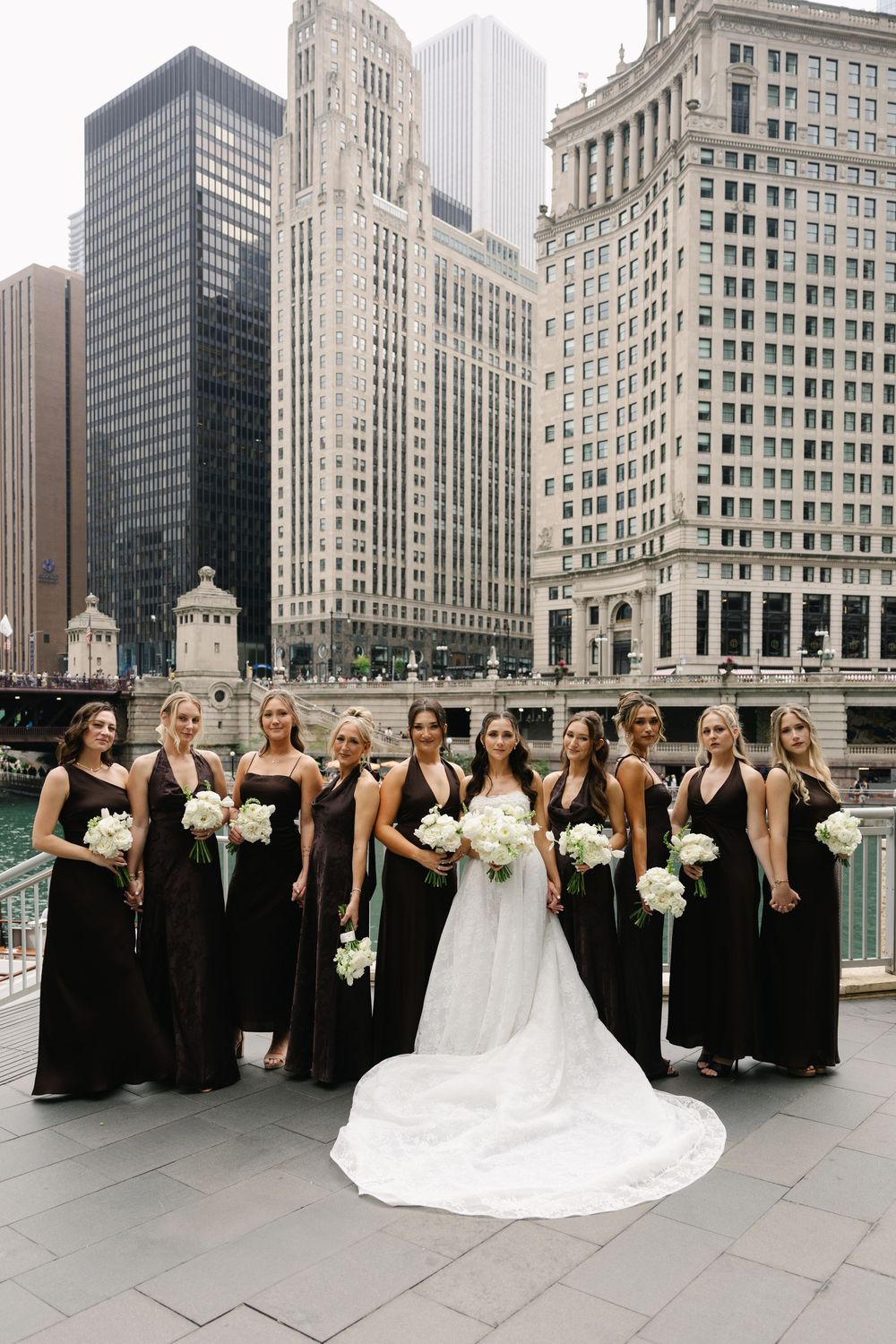 Bride in white gown with bridesmaids in black dresses holding white bouquets on Chicago Riverwalk with city skyline.