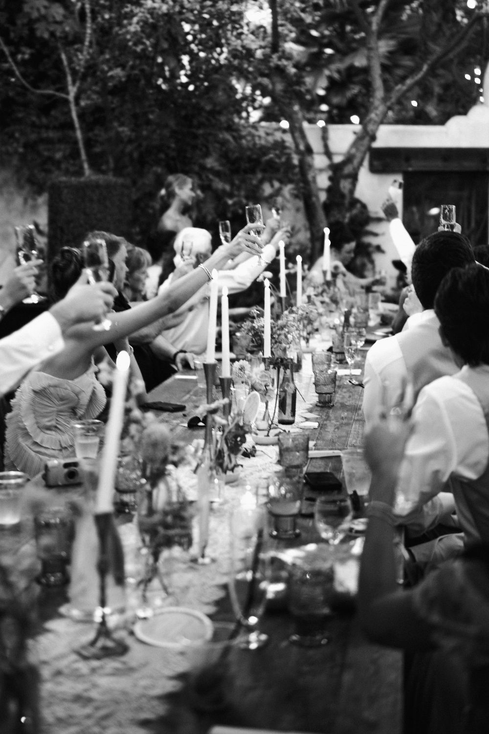 Black and white photo of wedding guests raising glasses in a toast at an elegant candlelit outdoor dinner reception.
