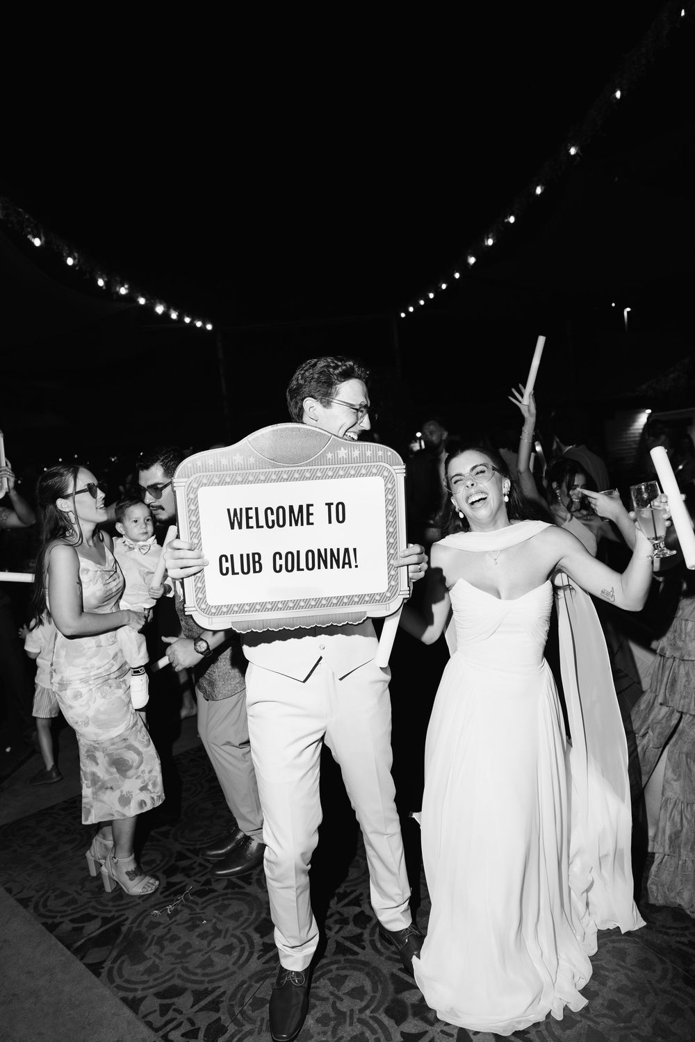 Bride and groom celebrate wedding reception holding Welcome to Club Colonna sign surrounded by cheering guests.