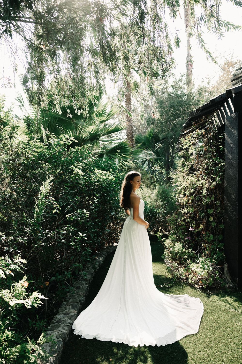 Bride in elegant white gown with long train stands in lush green garden surrounded by tropical trees and plants.
