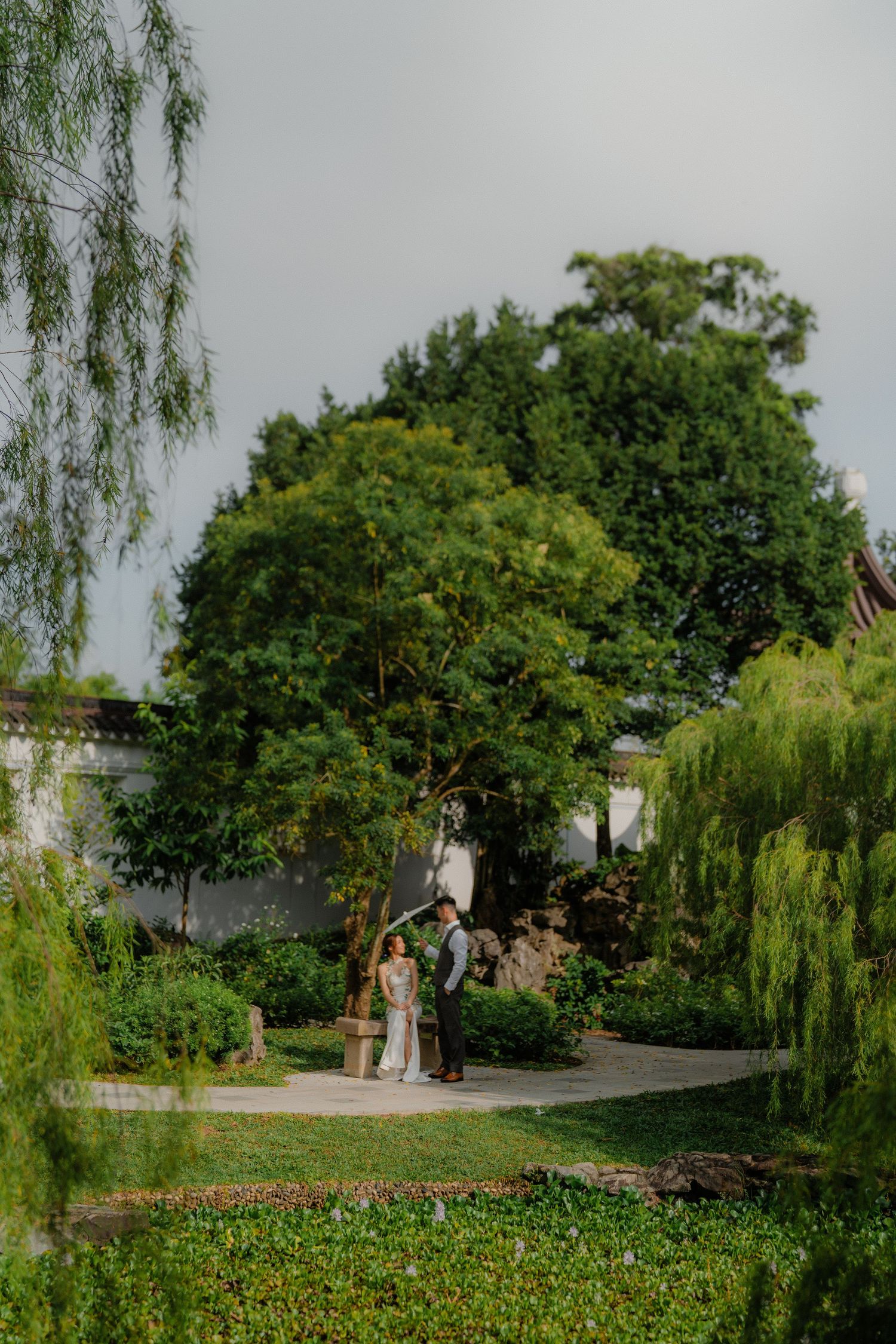Group gathers in a lush garden setting with weeping willow trees and blooming flowers on a summer afternoon.