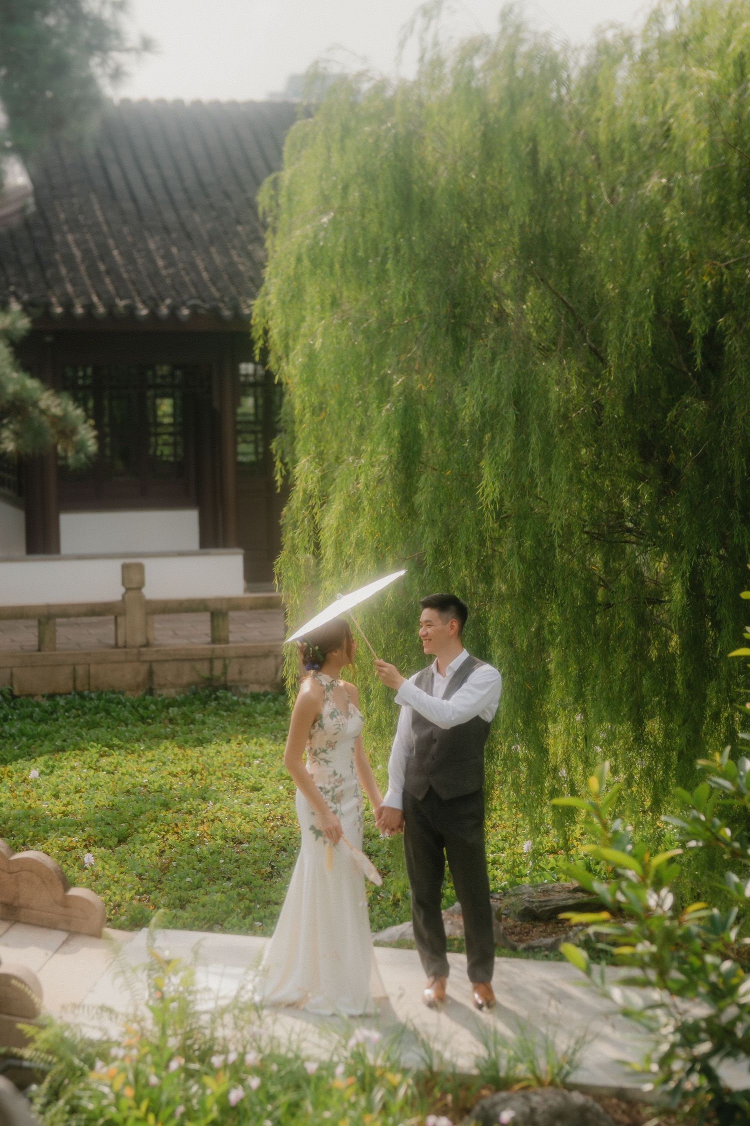 Wedding couple poses together amidst lush willow trees and traditional Chinese architecture.