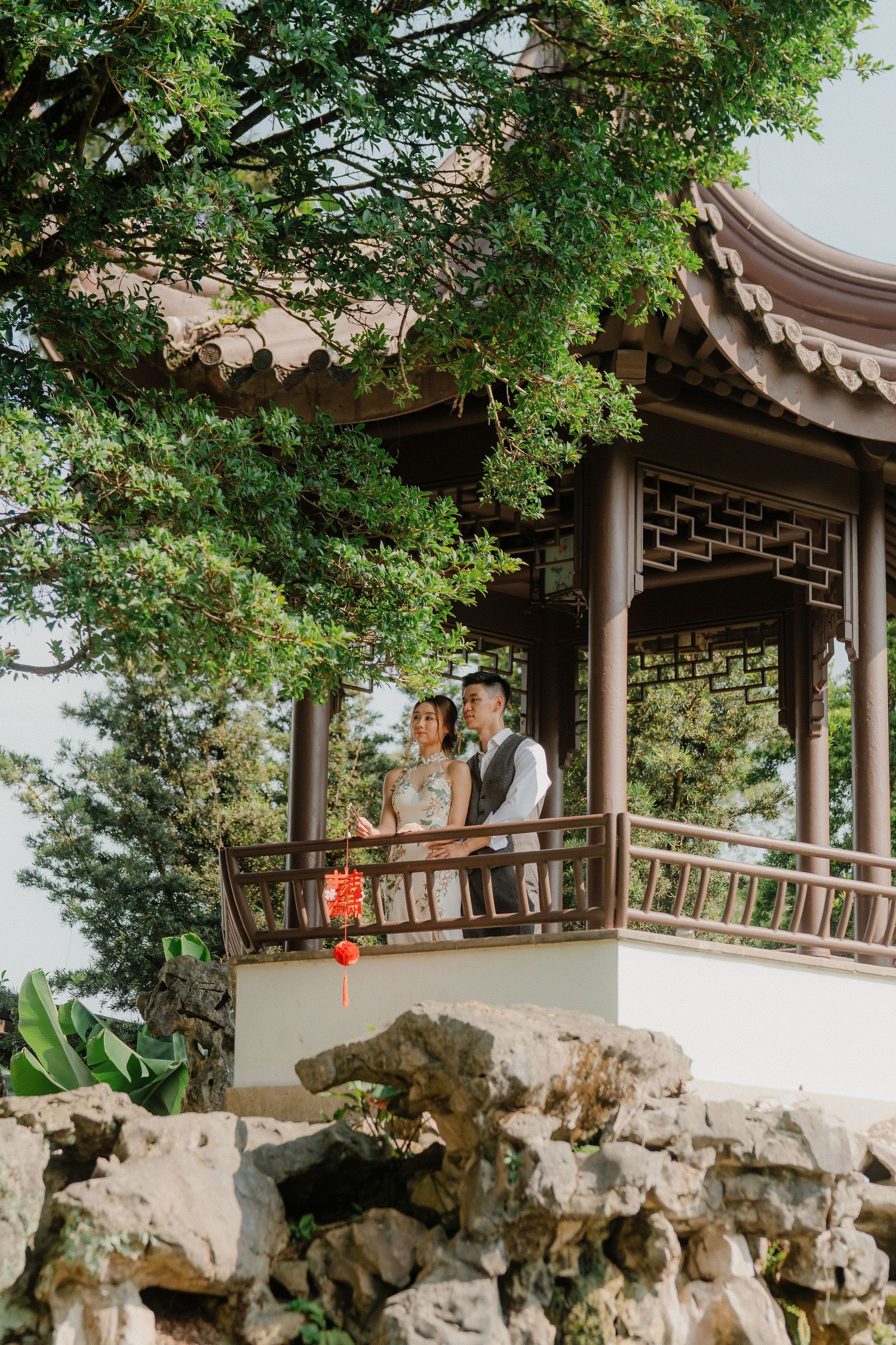 A couple shares a joyful moment on a traditional Asian pavilion surrounded by lush greenery and ornate stone features.