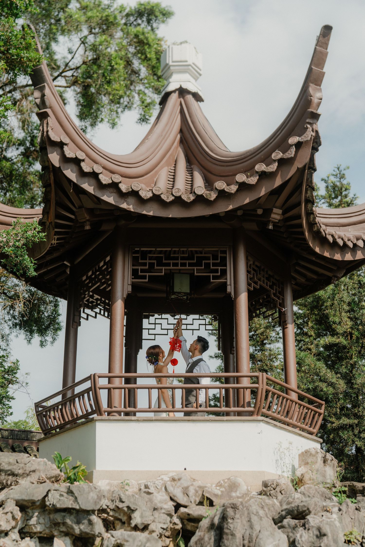 Traditional Chinese pagoda gazebo with curved roofline sits atop stone foundation in garden setting with green trees.