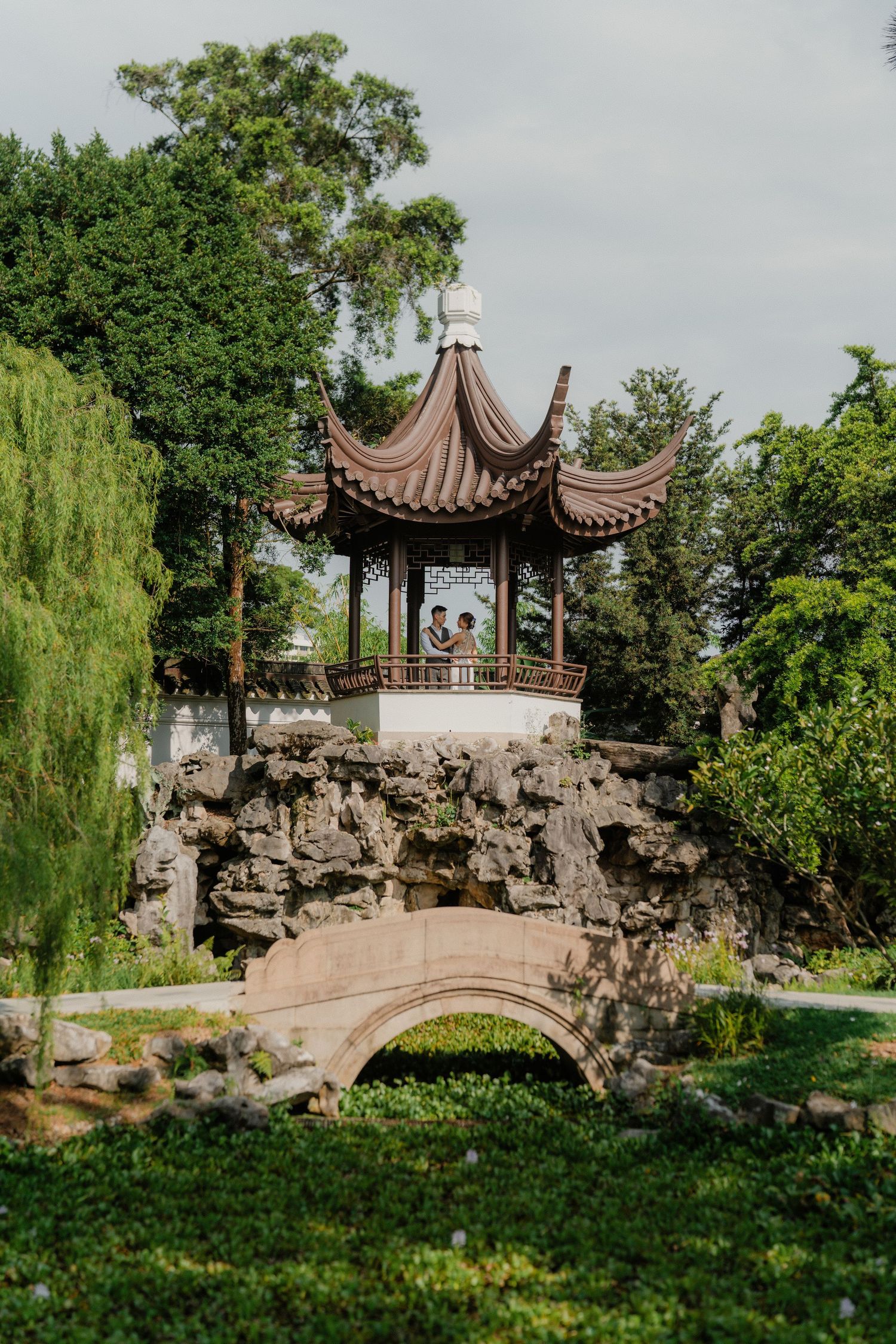 Traditional Chinese pagoda pavilion sits atop a decorative rock bridge in a peaceful garden with weeping willows.