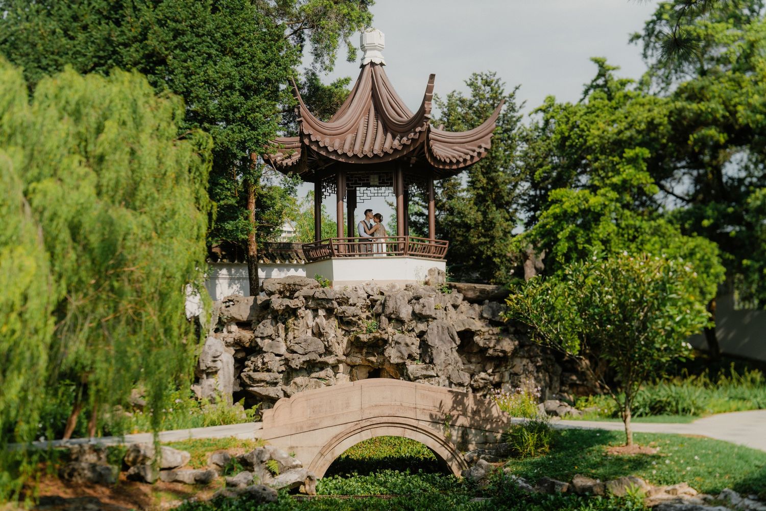 A scenic garden features traditional Chinese pavilions, bridges, and lush greenery in a tranquil landscape setting.