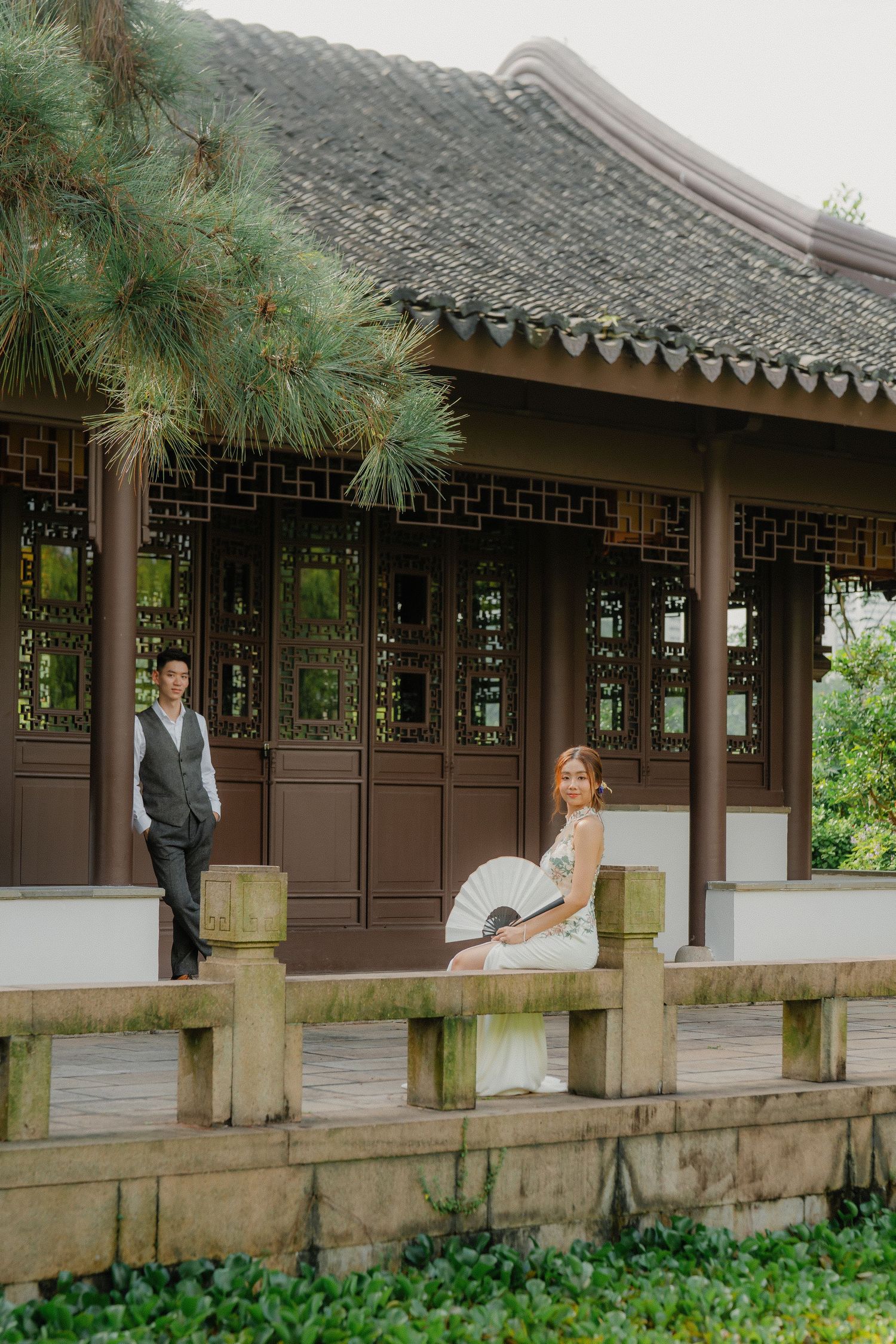 Traditional Chinese garden pavilion with tiled roof and ornate latticework details alongside a stone railing and lush greenery.
