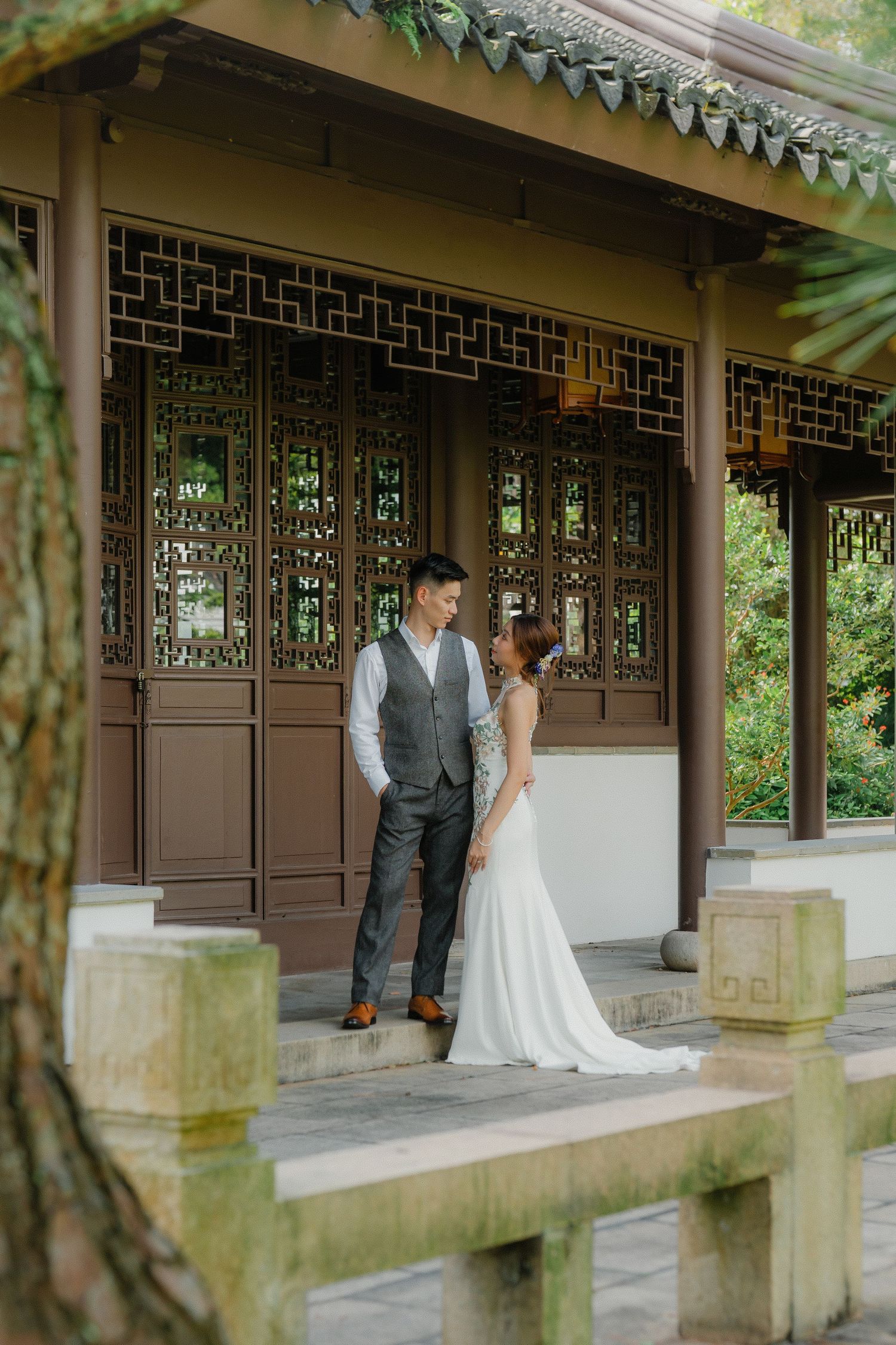 A couple stands together in a Chinese-style garden pavilion with ornate wooden lattice panels and traditional architecture.