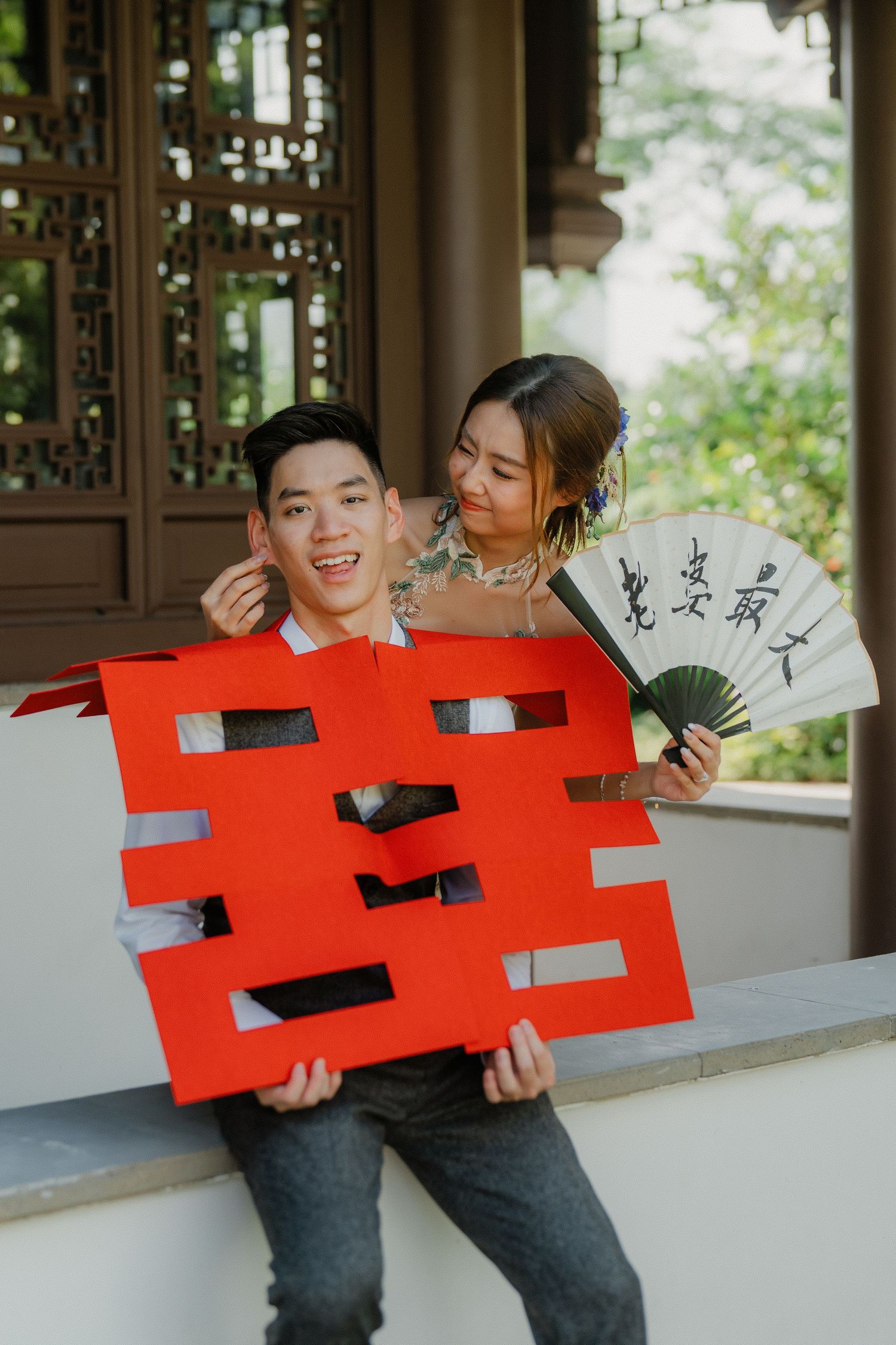 A couple poses playfully with traditional Chinese double happiness symbol and decorative fan.