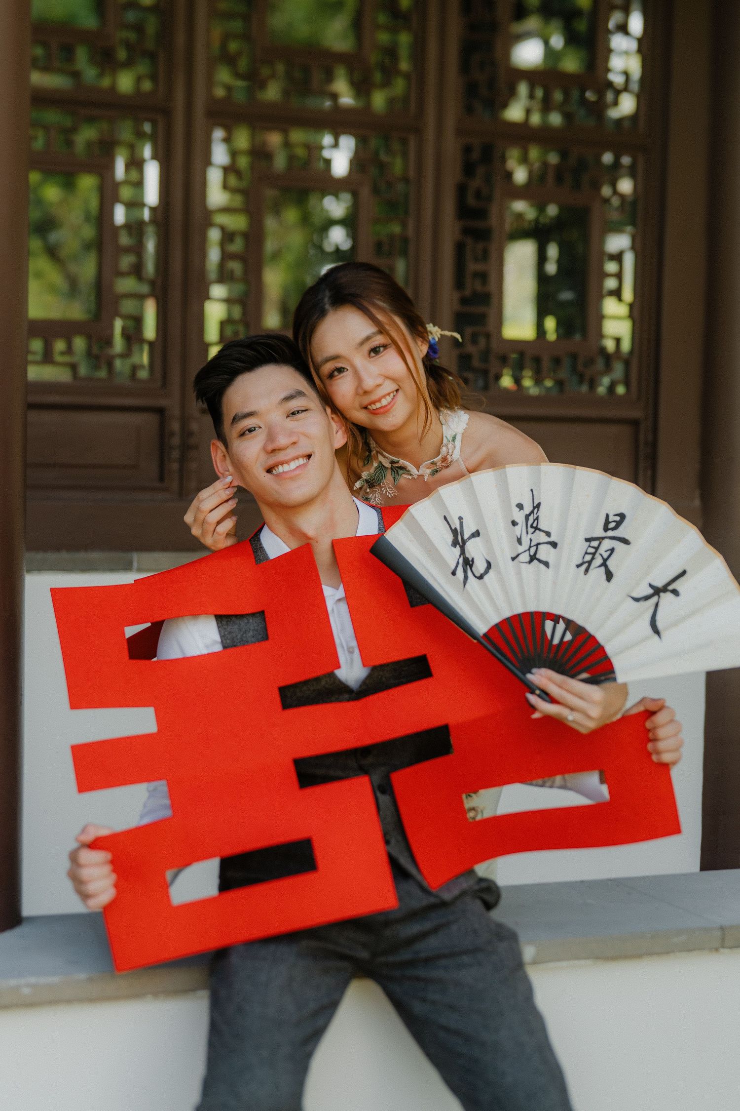 Young couple poses with traditional Chinese wedding decorations including a red double happiness symbol and calligraphy fan.