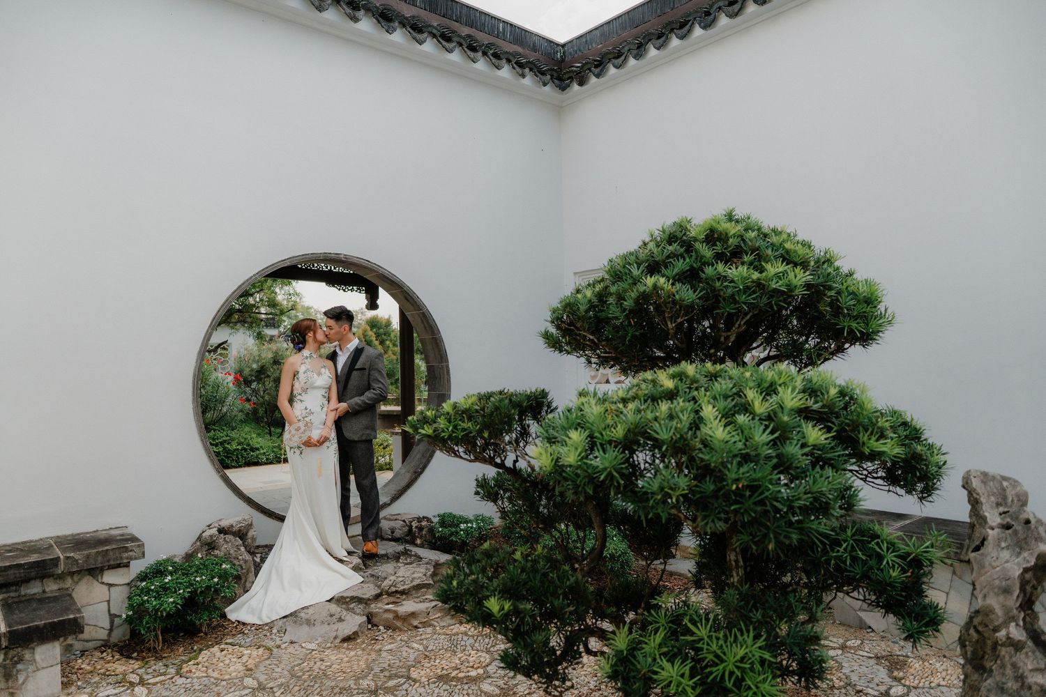 Couple stands in a traditional Chinese garden with a circular moon gate and shaped bonsai trees against white walls.
