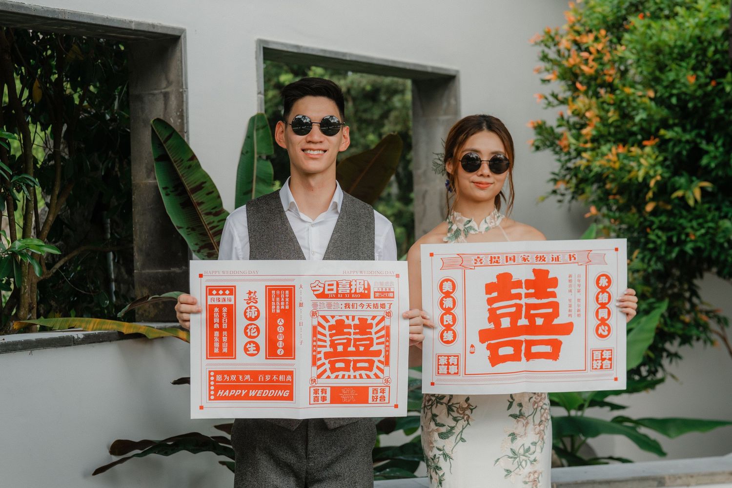 A couple in sunglasses holds traditional Chinese double happiness signs in a sunny garden setting.