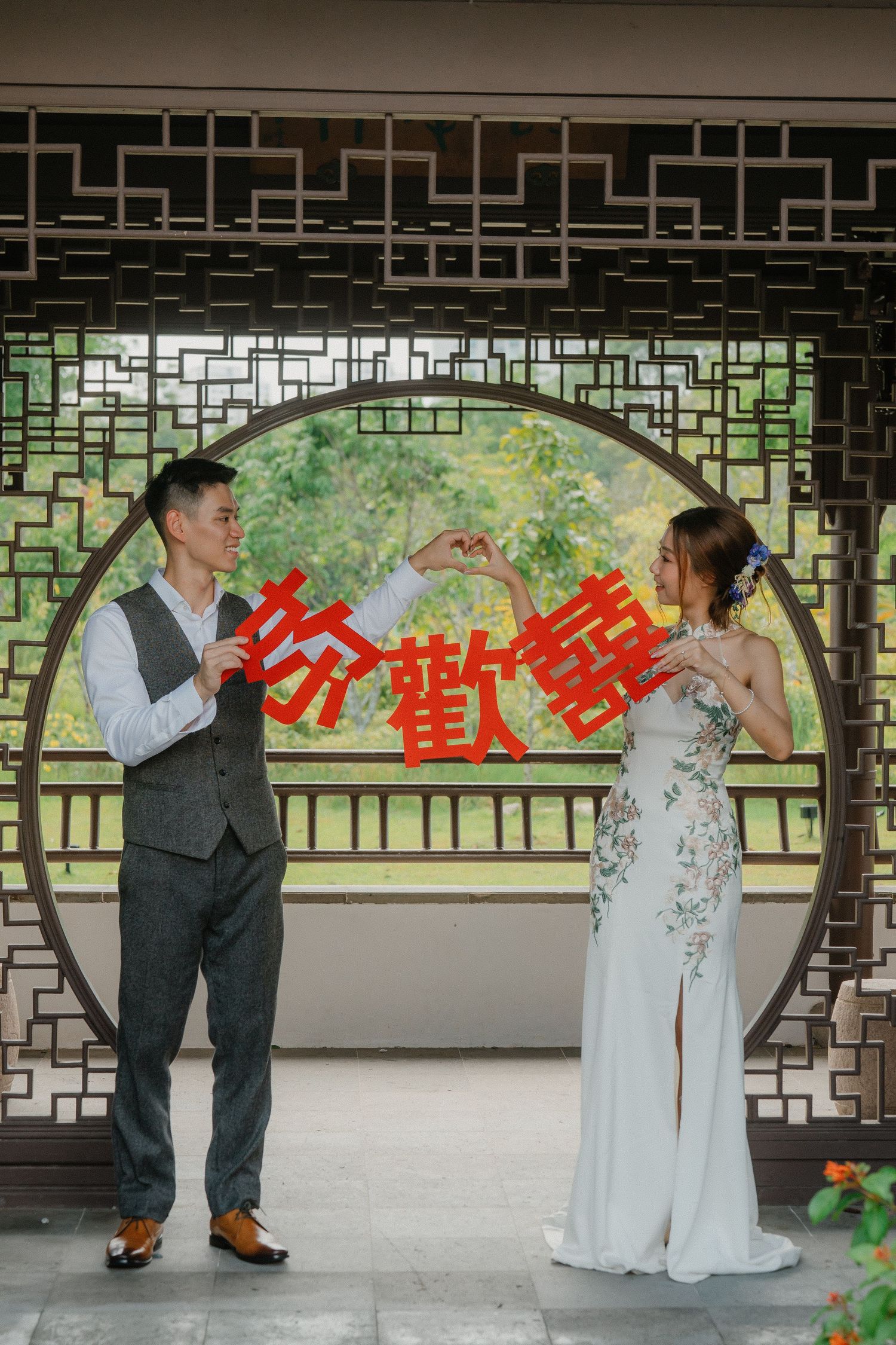 A couple poses in traditional Chinese wedding attire in front of a circular moon gate with red decorative characters.