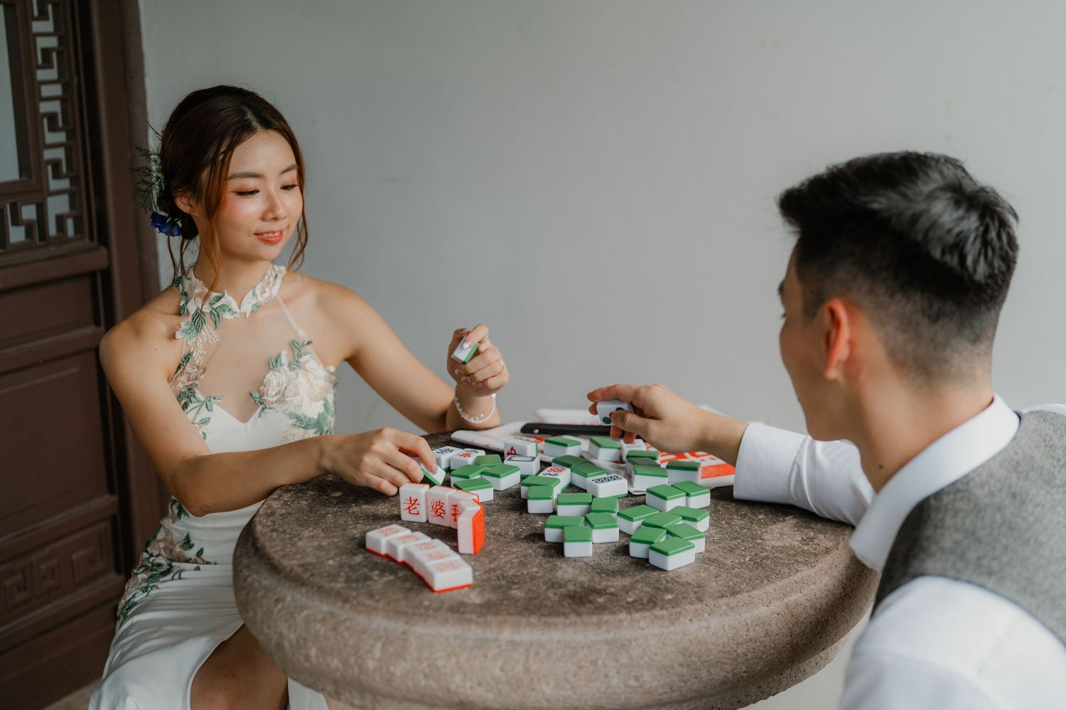Couple engaged in a game of mahjong at an ornate stone table in a traditional Chinese setting.