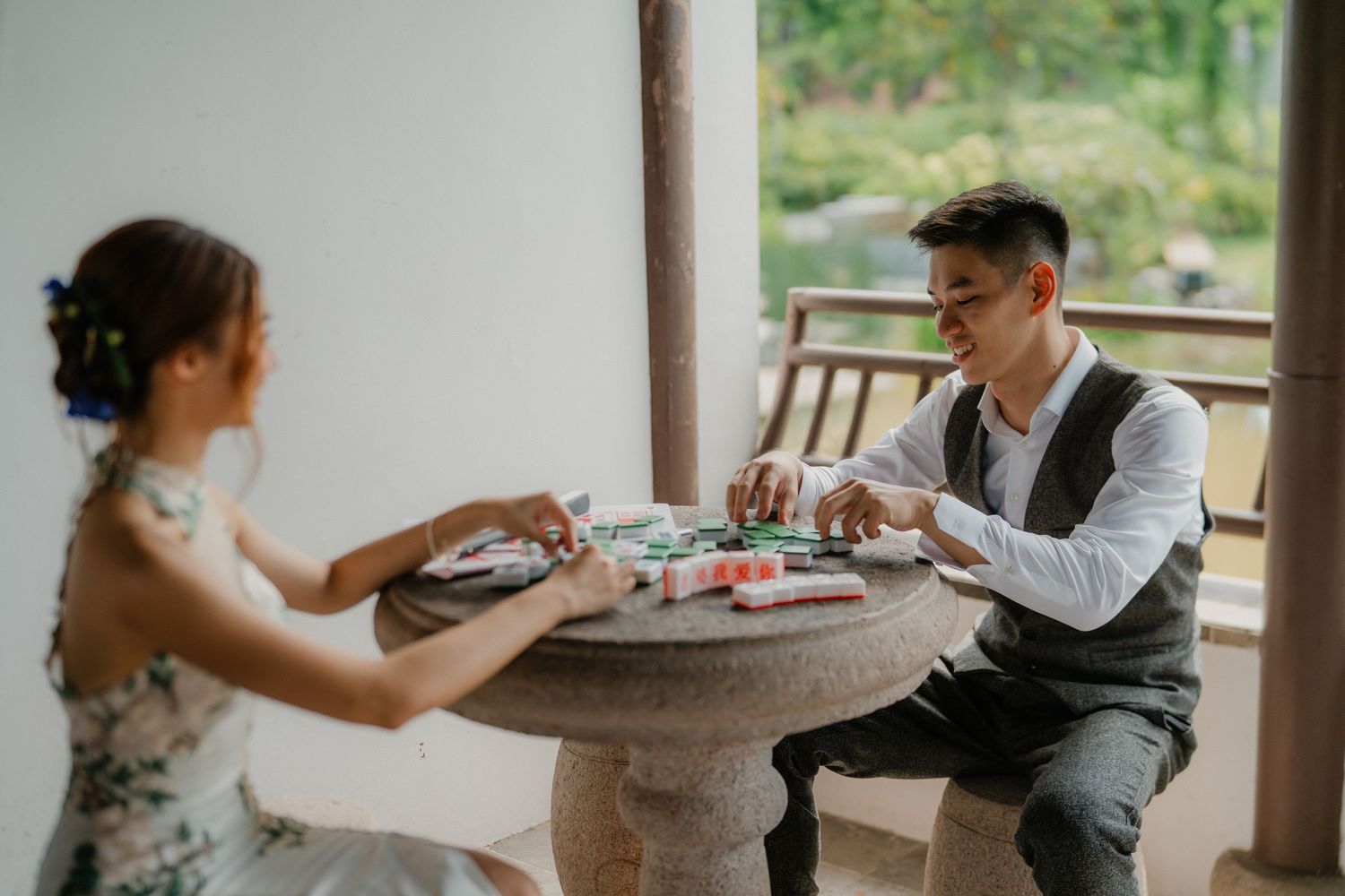 A couple shares a romantic moment while playing mahjong at a stone table on a porch.