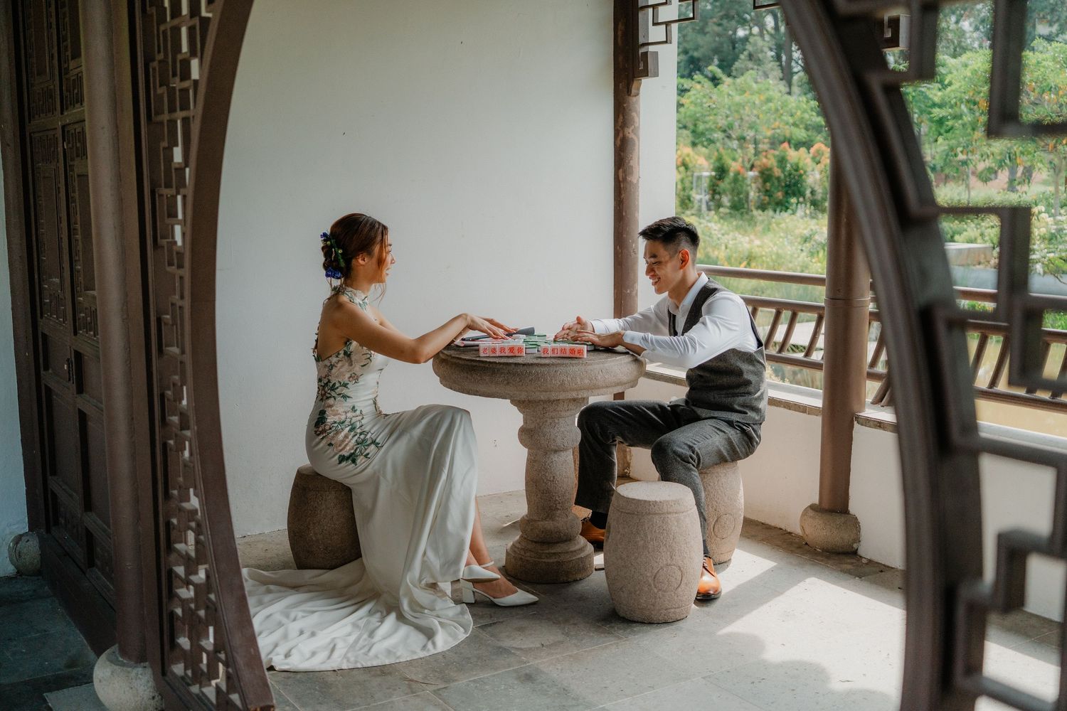 A couple shares a romantic moment playing cards at a round table on an ornate covered porch surrounded by natural scenery.