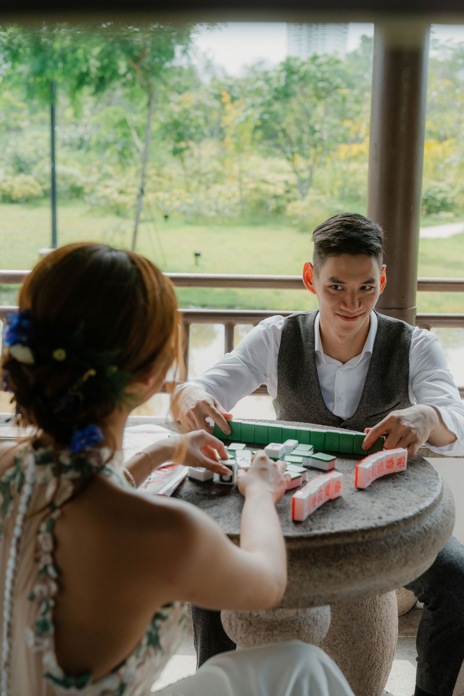 A couple playing with mahjong tiles on a round stone table outdoors.