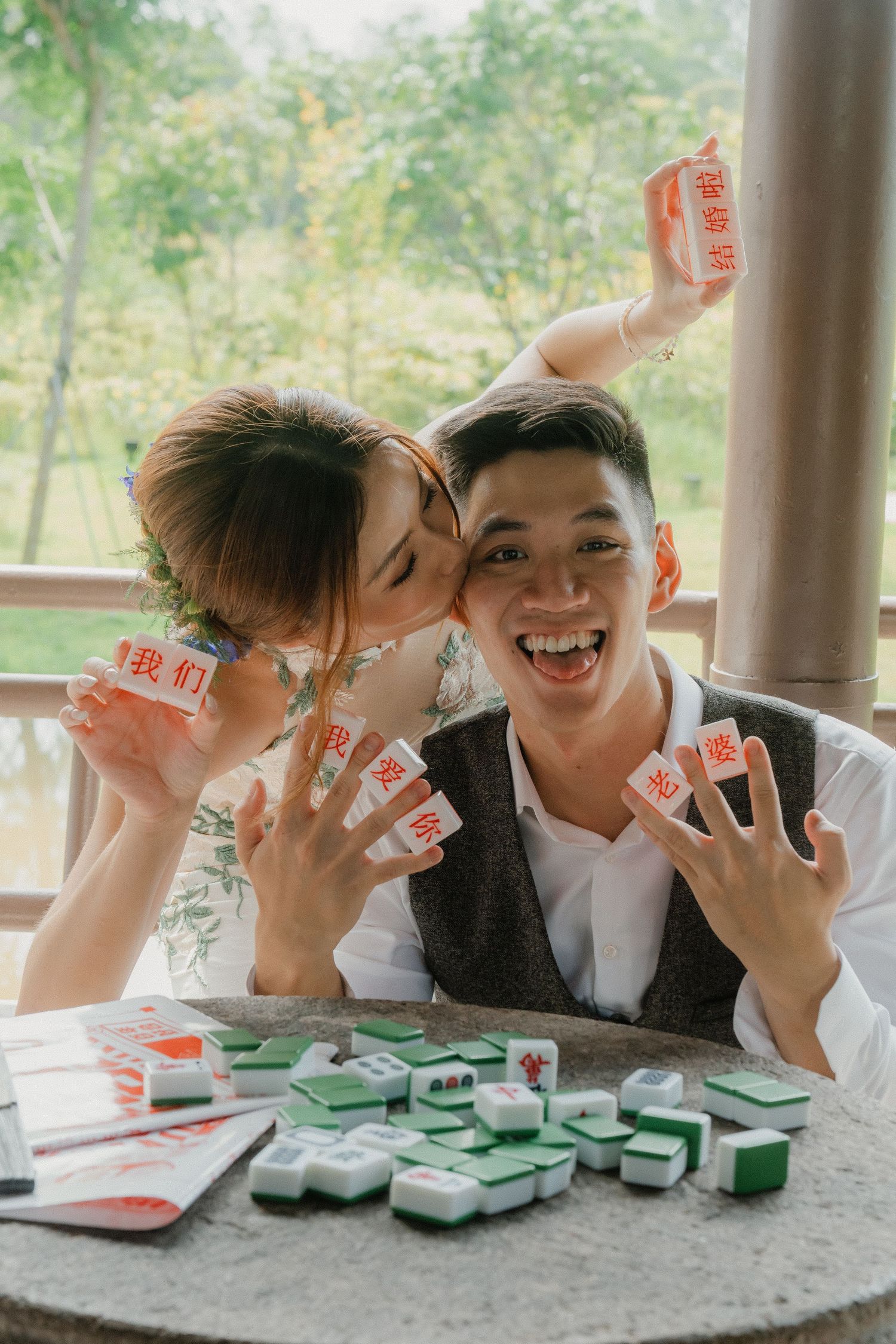 Playful couple showing playing cards while sitting at outdoor table with green card game pieces spread out.