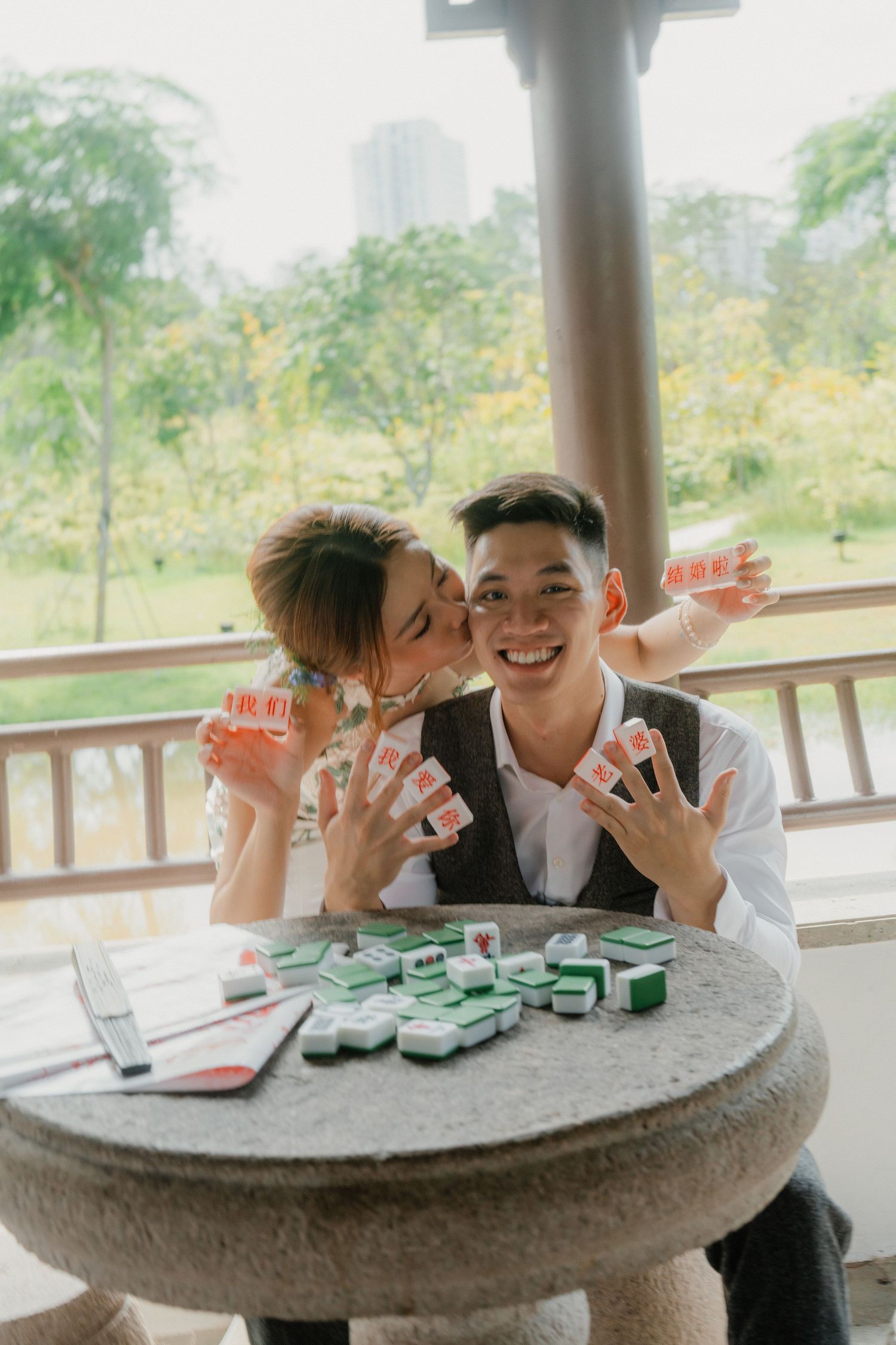 Two people arranging mahjong tiles playfully on an outdoor stone table.