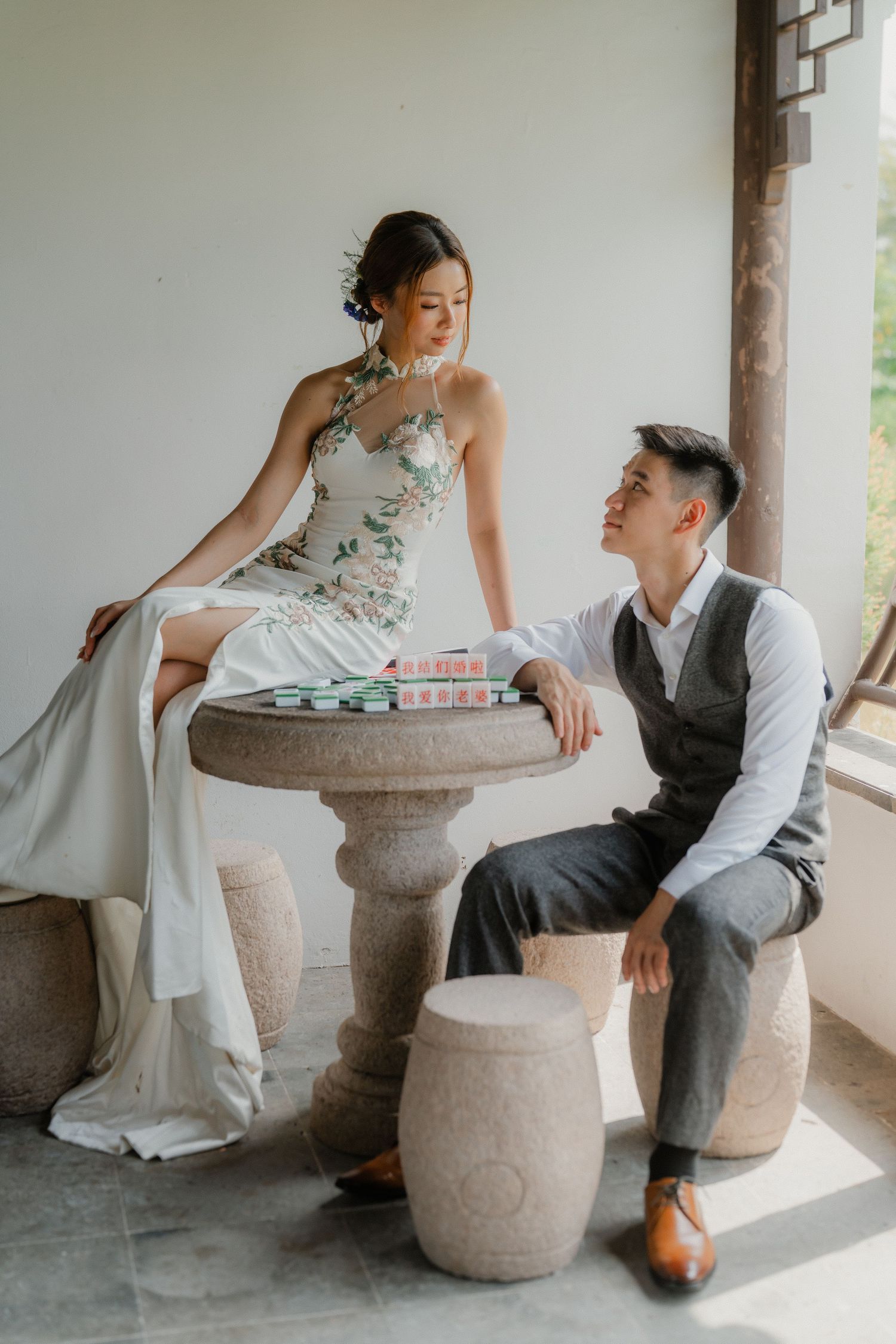 A romantic wedding photoshoot featuring a couple posing playfully around a stone table on a covered porch.